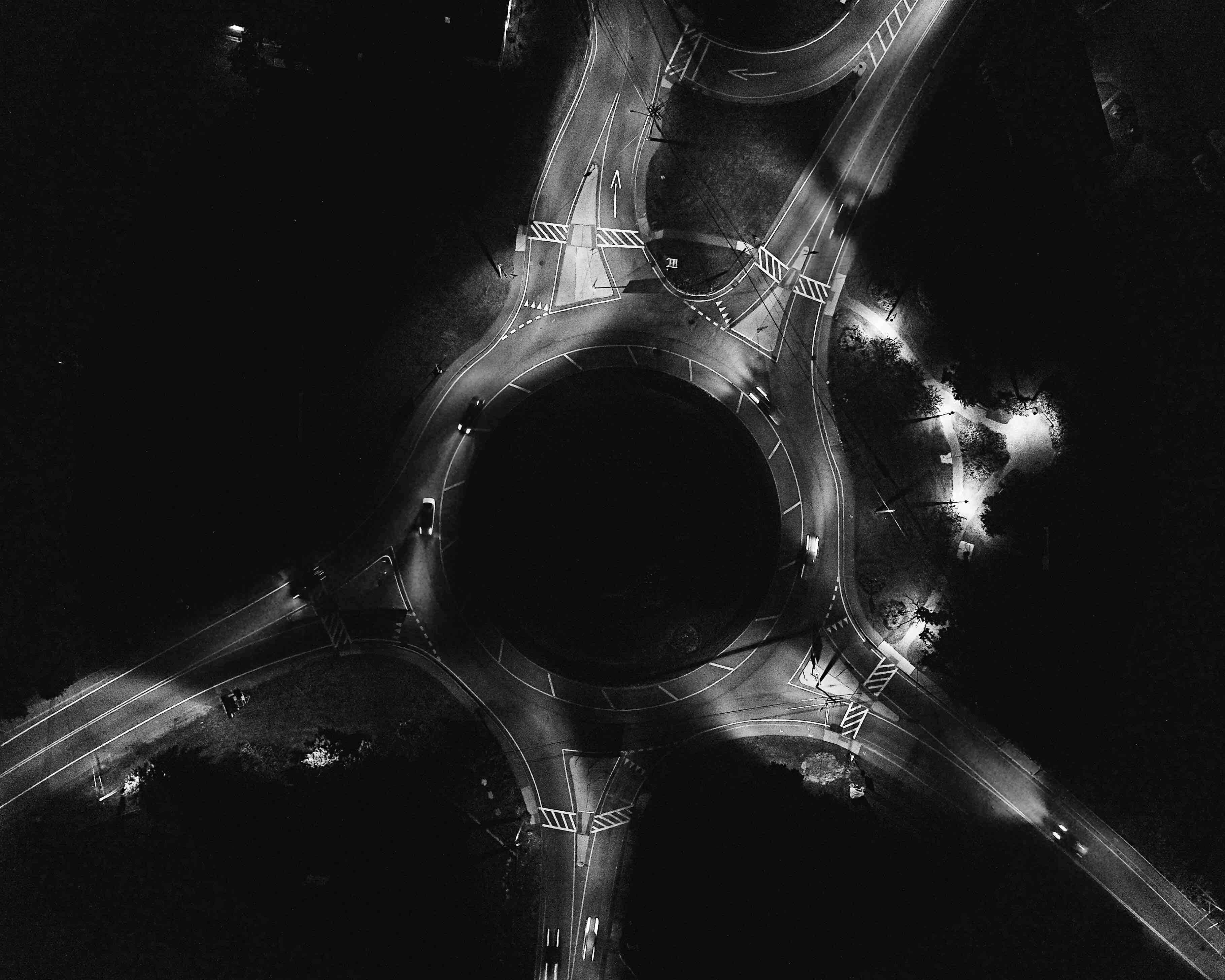 Dark Crossroad 桌布 壁紙 — 從空中拍攝的圓形道路交叉口的黑白夜景，帶有發光的車頭燈和路燈。