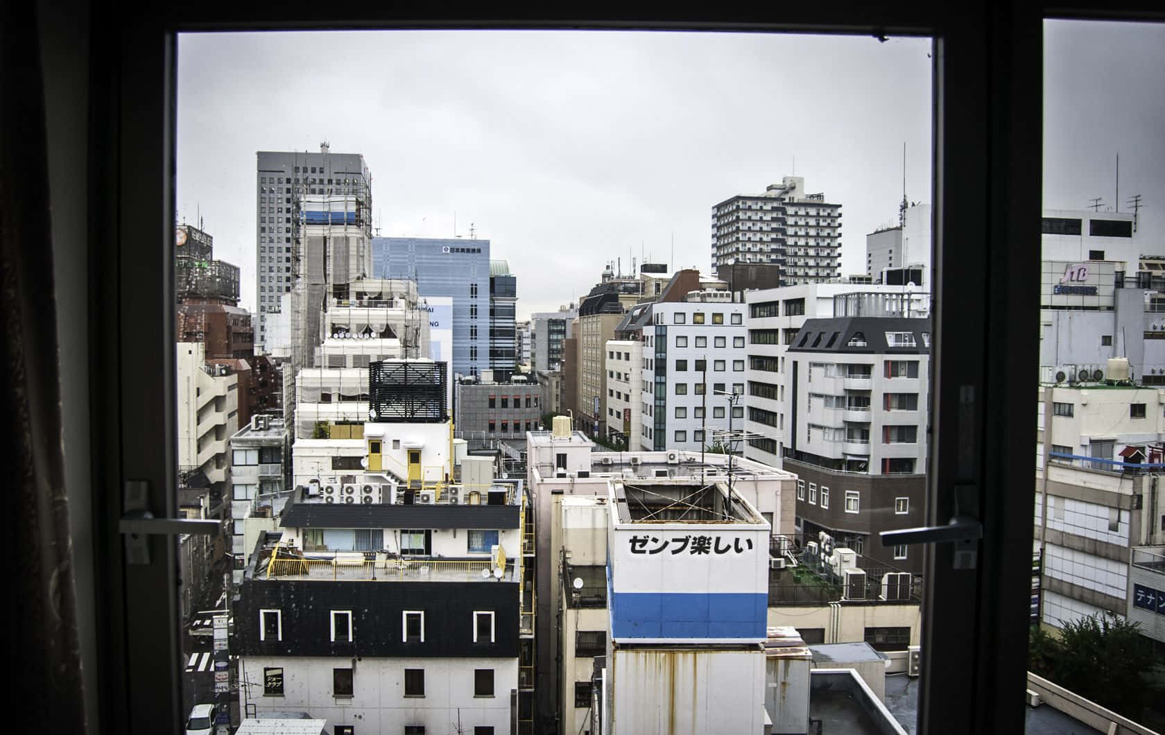 日式審美桌面 桌布 壁紙 — 從窗戶望出去，框住了東京密集的城市天際線，天空有灰色雲層，一棟建築物上有 zenpureshii 標誌。