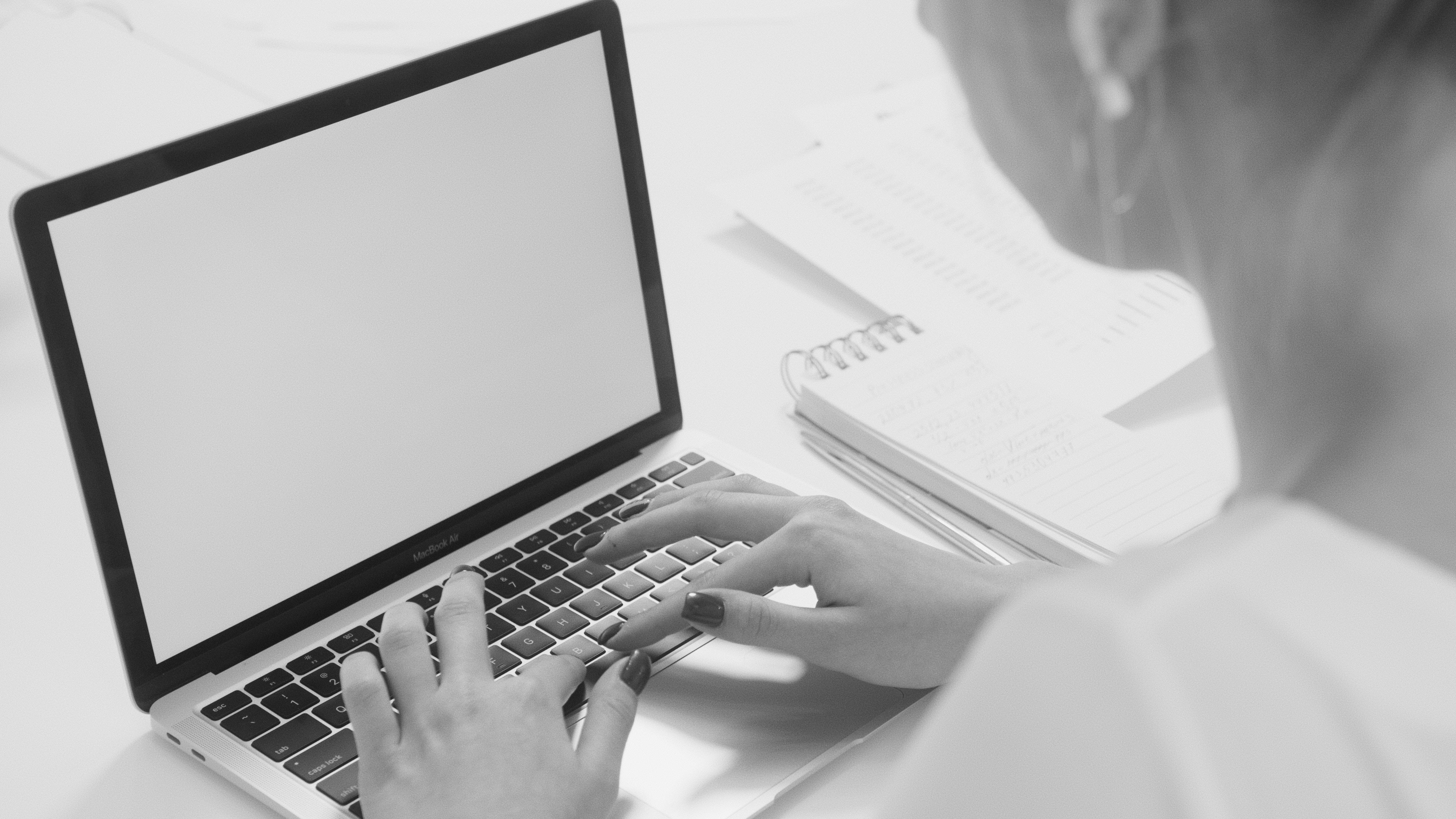 aesthetic tumblr laptop 桌布 壁紙 — 從肩膀上方拍攝的黑白照片，一個人在空白螢幕的筆記型電腦上打字，旁邊有一個螺旋筆記本。