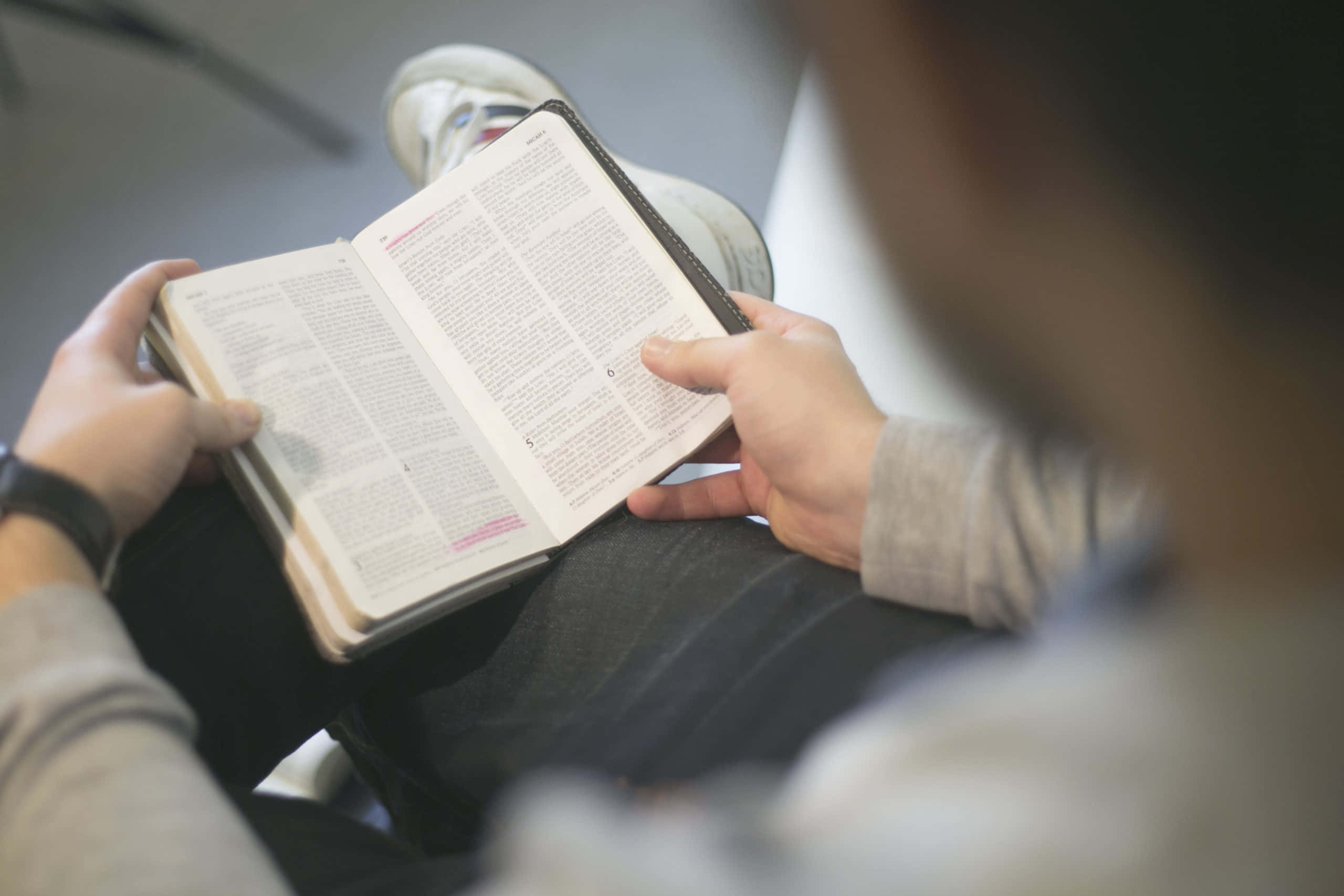 聖經 桌布 壁紙 — 從肩膀上方看到一個人穿著運動鞋坐著閱讀一本厚書。