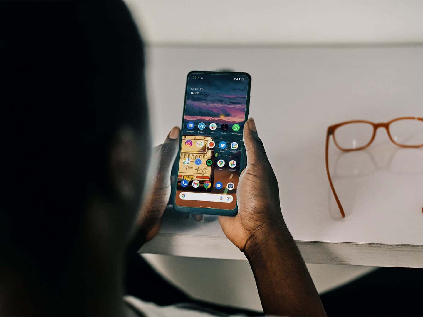 Smartphone 桌布 壁紙 — 從肩膀上方觀看，雙手拿著智慧型手機，顯示色彩繽紛的應用程式網格，背景是充滿活力的日落桌布。