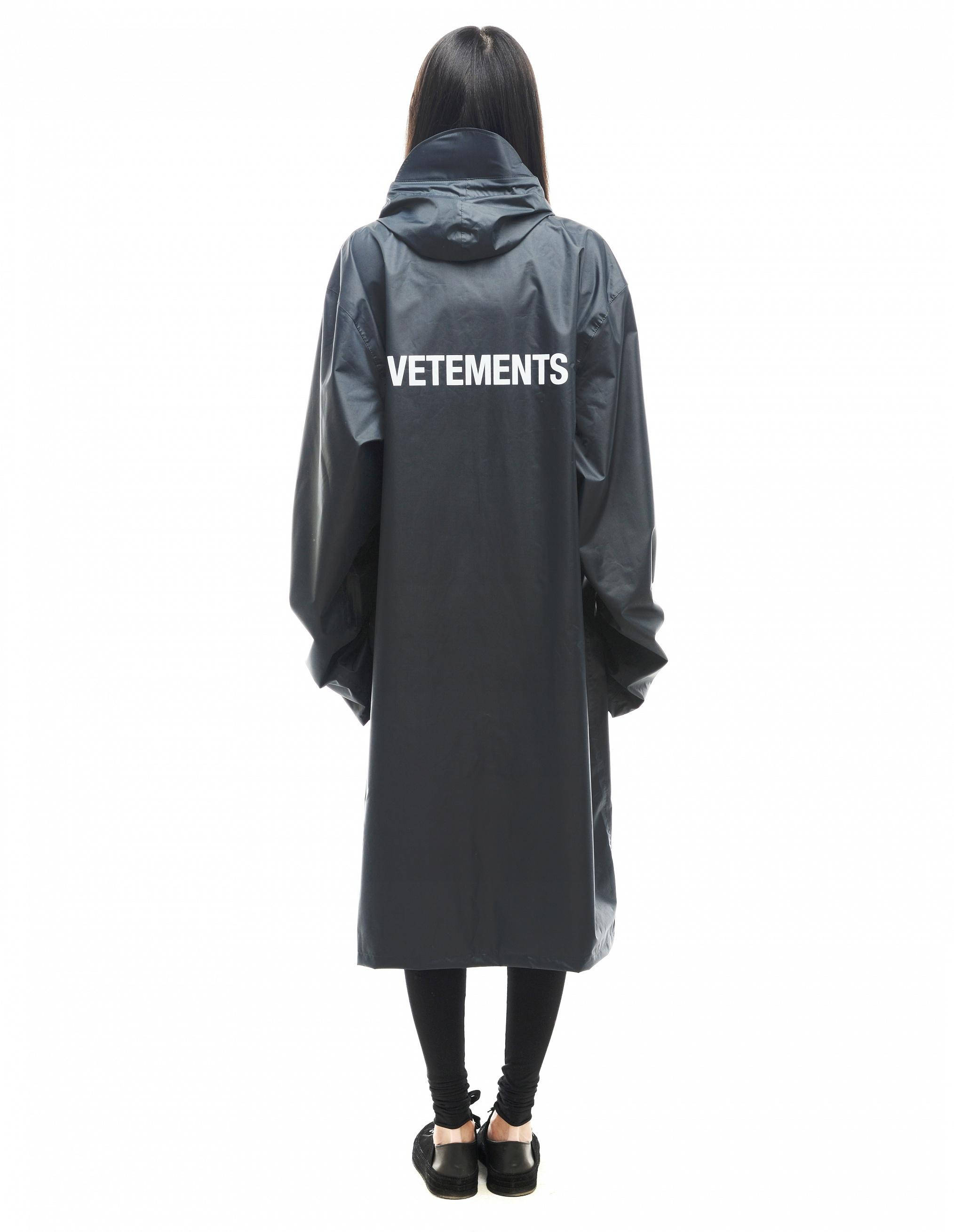 Vetements 桌布 壁紙 — 從背後看到的一個人穿著一件長黑色連帽雨衣，背面有醒目的白色VETEMENTS品牌標誌。