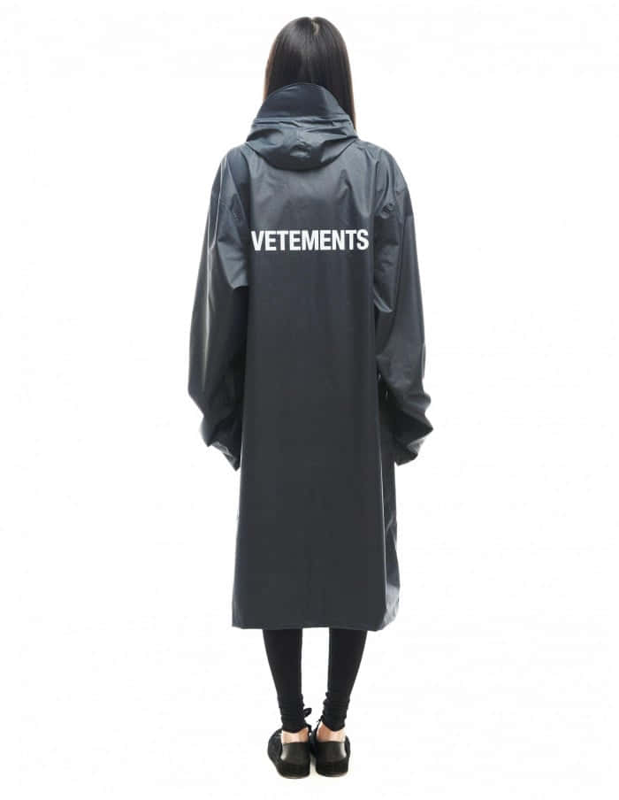 vetements 桌布 壁紙 — 從背後看到的人穿著一件長黑色雨衣，背面有白色VETEMENTS文字，背景為純白色。
