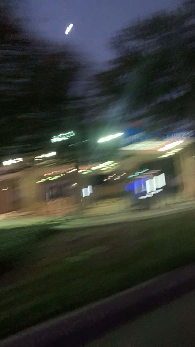 Building 桌布 壁紙 — 從行駛中的車輛看到的條紋夜景，顯示在黑暗天空下模糊的路燈、樹木和商店門面。