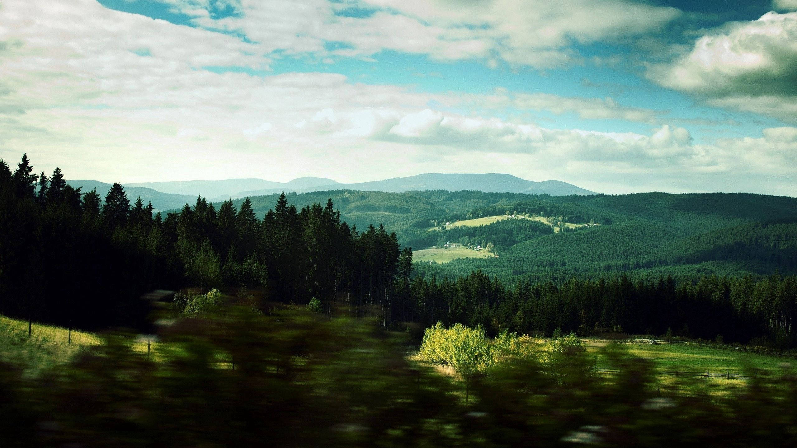 風景 桌布 壁紙 — 從行駛中的車輛看到的風景，展現了起伏的綠色山丘、森林和遠處的山脈，天空部分多雲。
