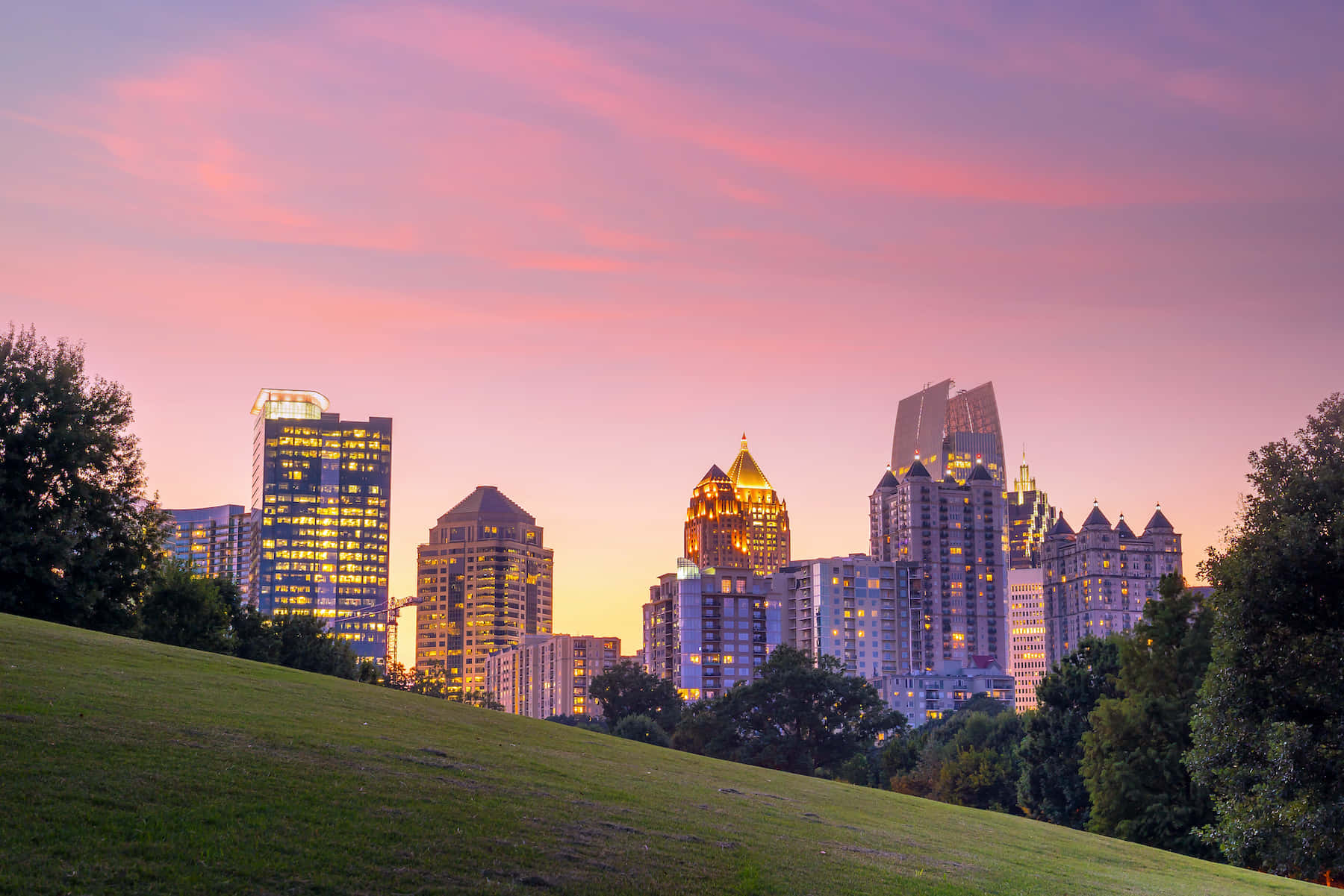 Atlanta Georgia 桌布 壁紙 — 從長滿草的山丘上觀賞，黃昏時分充滿活力的城市天際線，閃爍的摩天大樓映襯著粉紅色和紫色的天空。