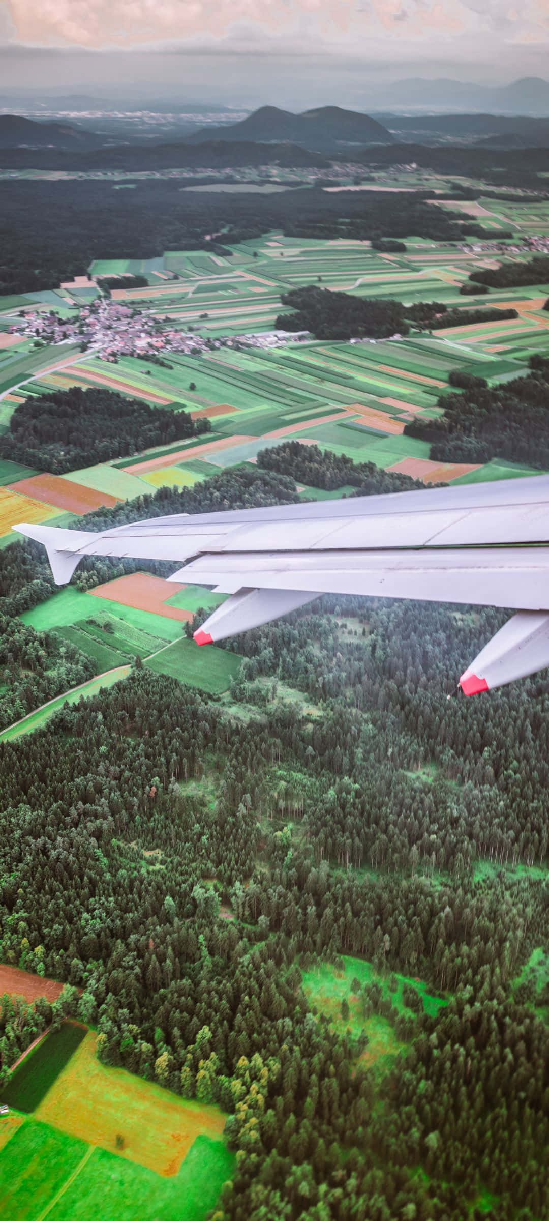 1080x2400 桌布 壁紙 — 從飛機窗戶俯瞰，機翼下方是拼布般的綠色田野、森林和遠山，天空多雲。