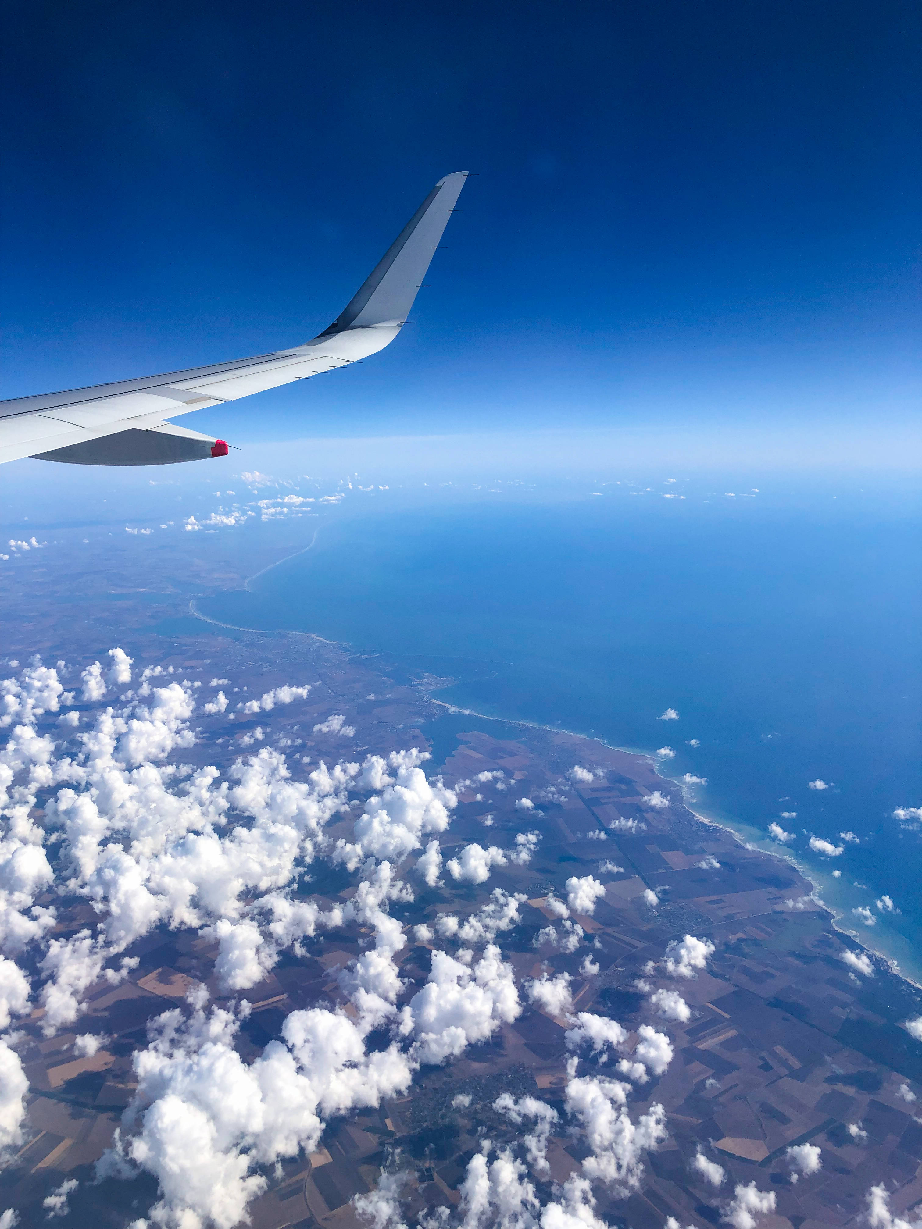 飛機 桌布 壁紙 — 從飛機窗戶俯瞰，機翼在蓬鬆雲層之上，下方是海岸景觀，天空清澈湛藍。