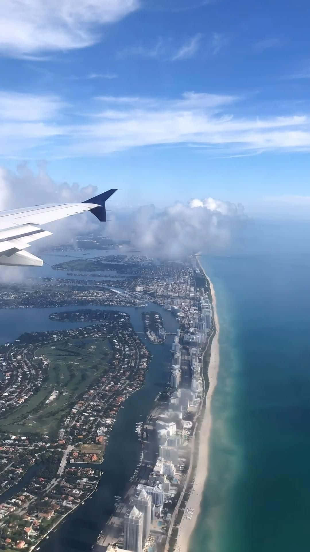 Travelling 桌布 壁紙 — 從飛機窗戶俯瞰海岸城市景觀，包括沙灘、海洋和散落的雲朵，在明亮的藍天下。