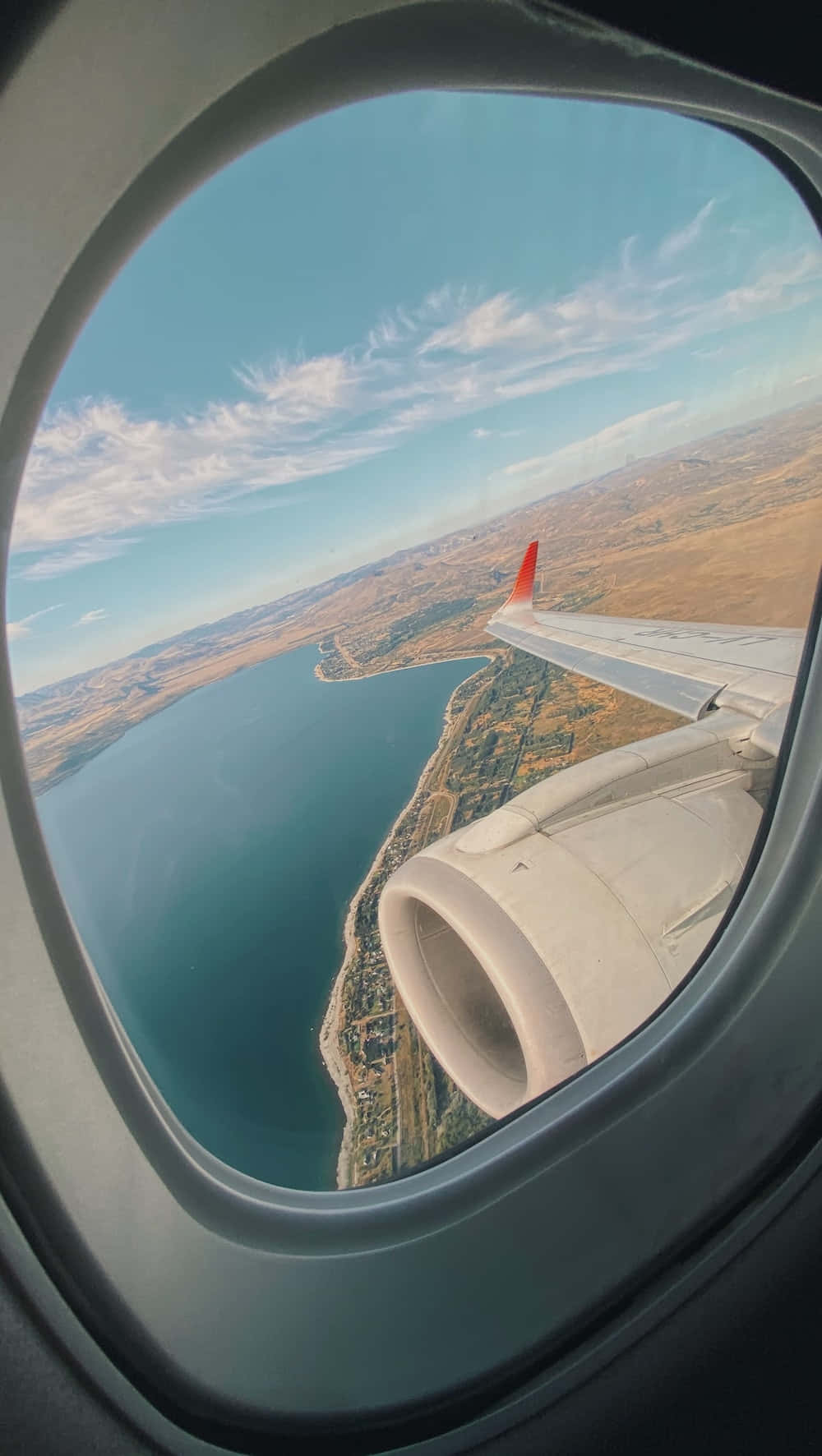 Traveling 桌布 壁紙 — 從飛機窗戶俯瞰的航拍圖，顯示機翼引擎在清澈天空下，位於藍色海水和棕色地形的海岸景觀之上。
