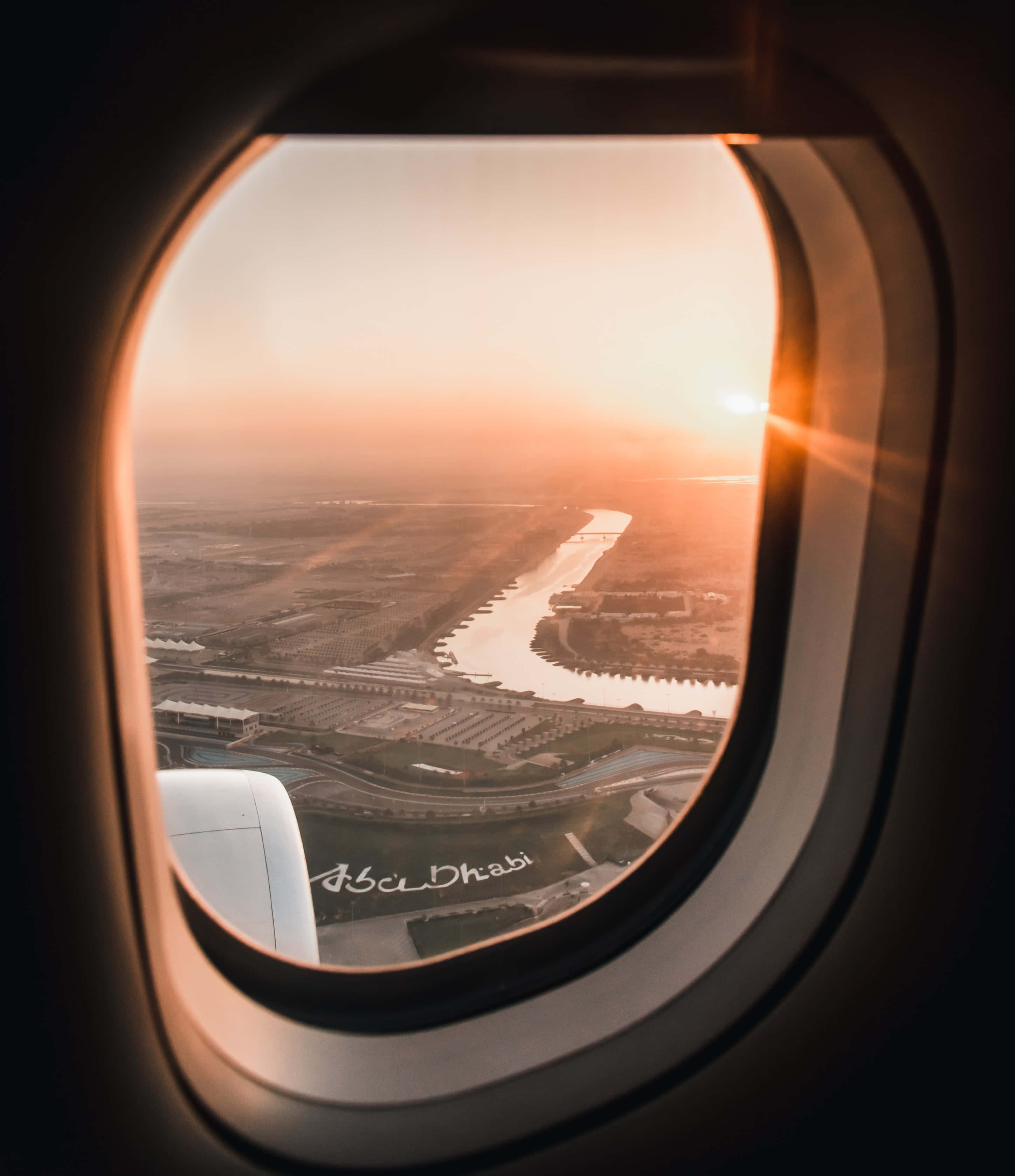 Airplane Window 桌布 壁紙 — 從飛機窗戶俯瞰阿布達比日落時分，金色光芒照亮下方的河流與城市景觀。