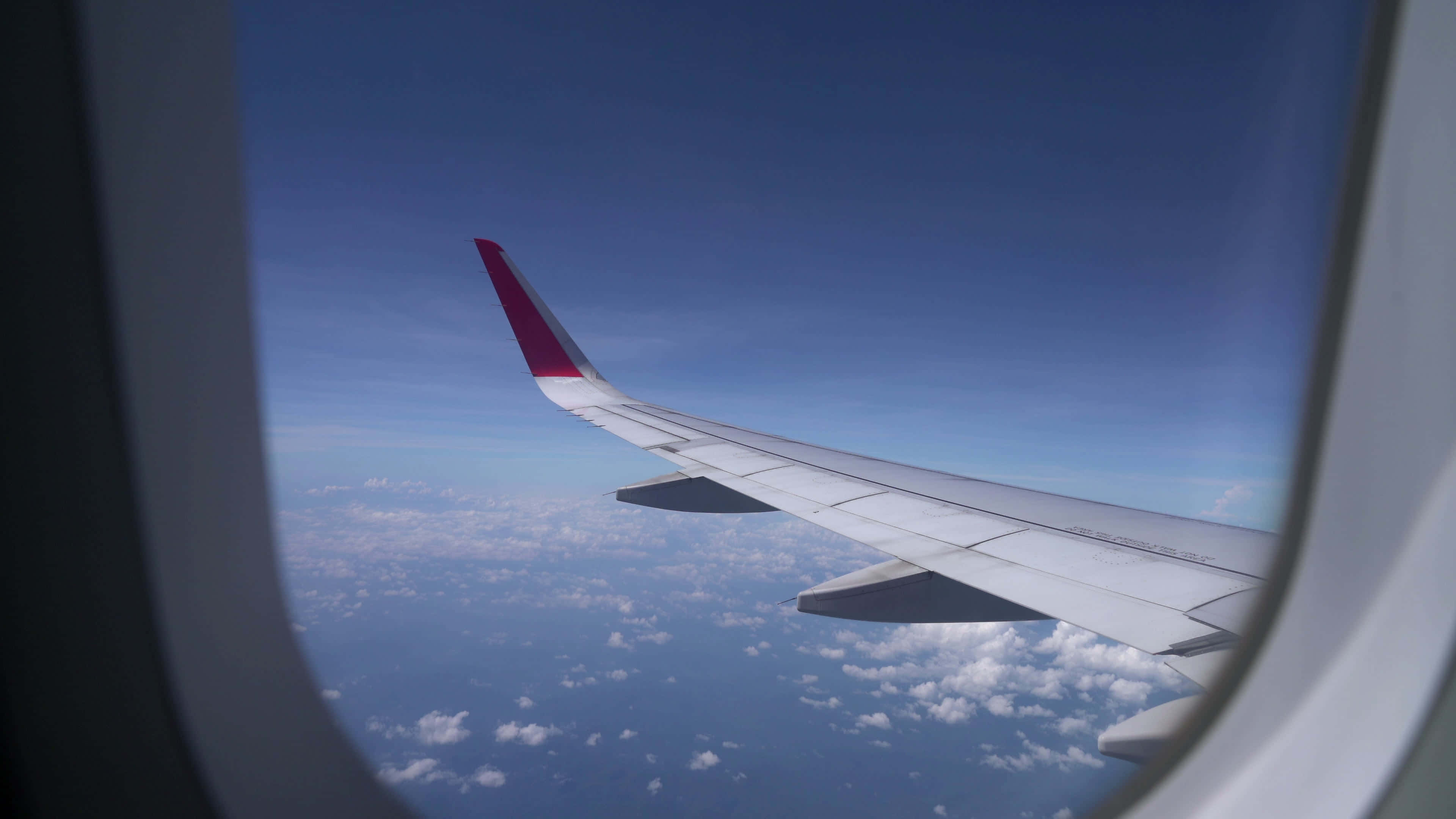 Airplane Window 桌布 壁紙 — 從飛機窗戶看到的紅色尖端飛機機翼，位於蓬鬆白雲之上，清澈藍天之下。