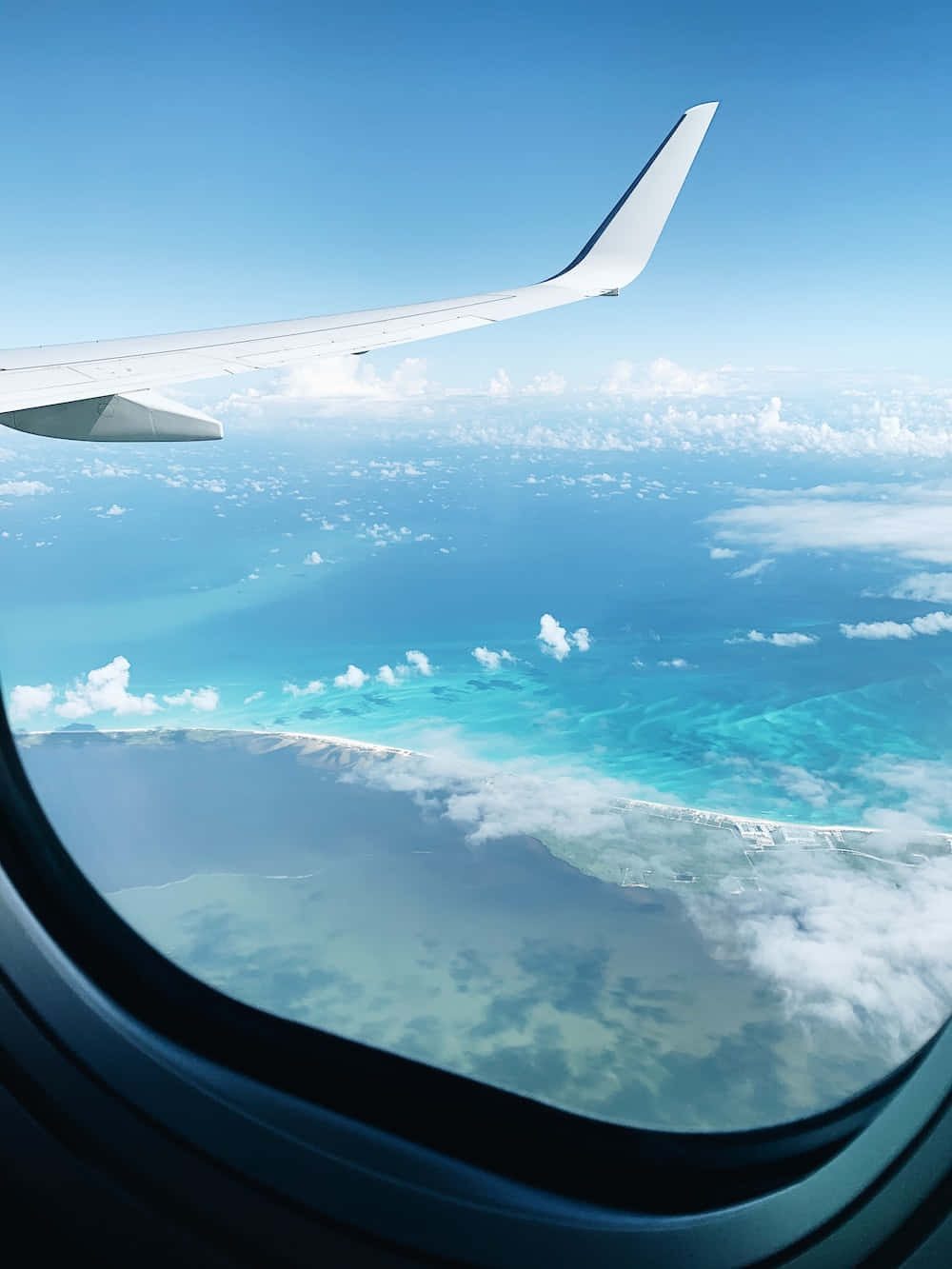 Airplane Window 桌布 壁紙 — 從飛機窗戶看到的飛機機翼，俯瞰碧綠海洋和散落的雲朵，在清澈的藍天下。