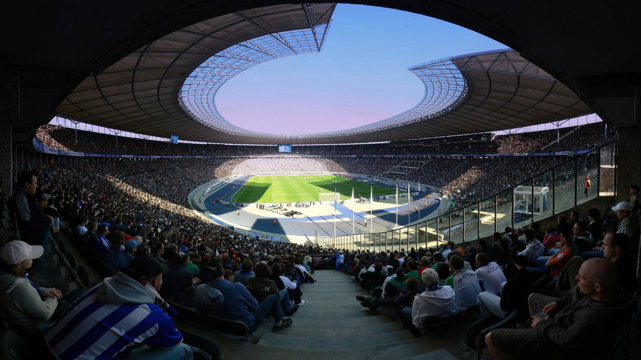 Stadium 桌布 壁紙 — 從體育場座位全景視角俯瞰擠滿觀眾的競技場，綠色場地和跑道在暮色天空下。