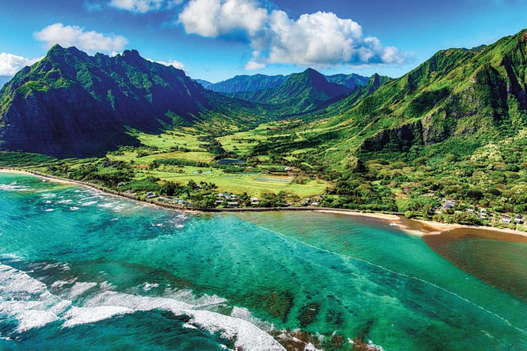 Hawaiian Island 桌布 壁紙 — 從高空俯瞰，一個充滿活力的綠松石色海灣坐落在鬱鬱蔥蔥的綠色山脈之間，上方是明亮的藍天和蓬鬆的白雲。