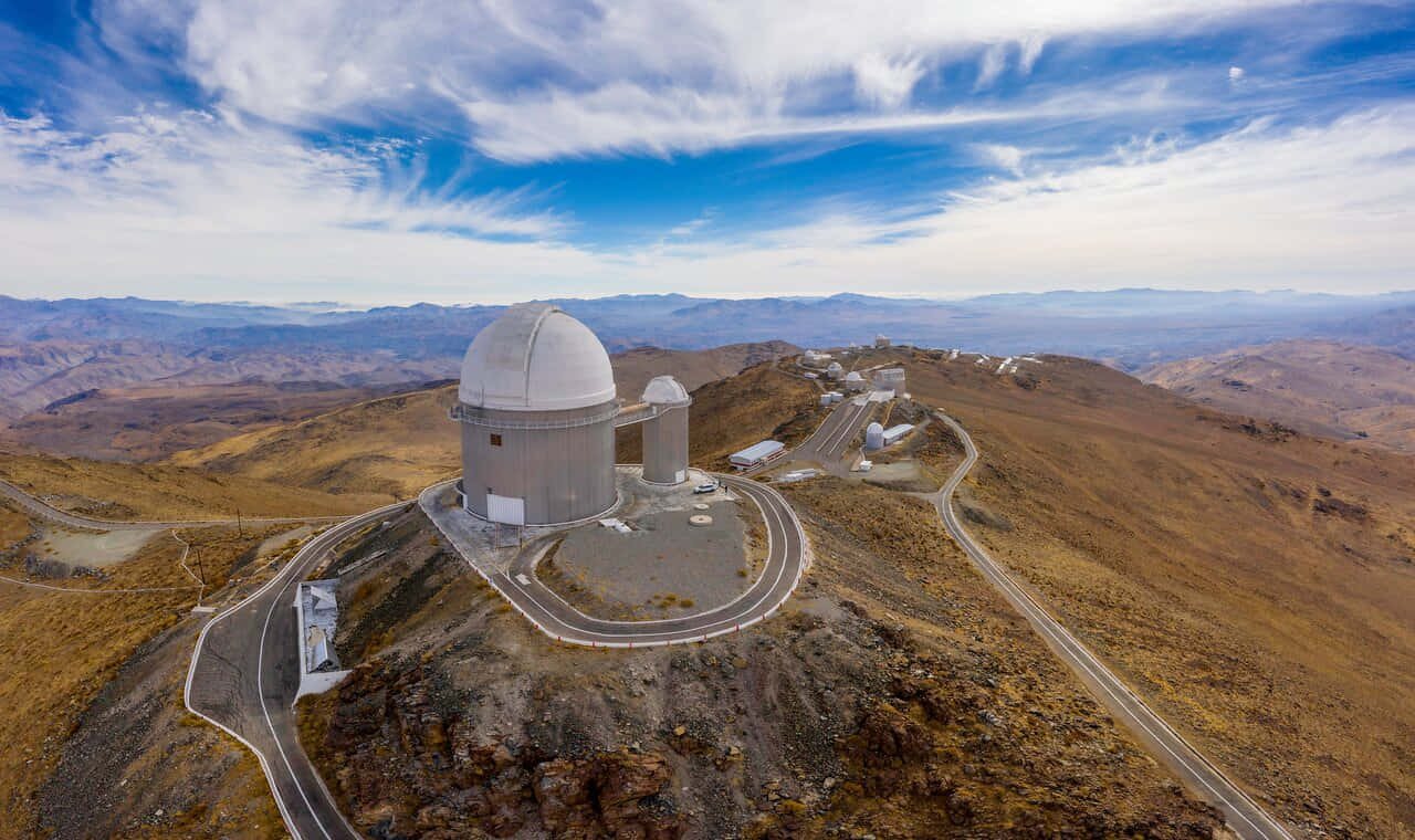 Observatory 桌布 壁紙 — 從高空俯瞰，一個大型天文台圓頂坐落於荒蕪的山頂上，蜿蜒的道路和遠處的山脈在藍天白雲下清晰可見。