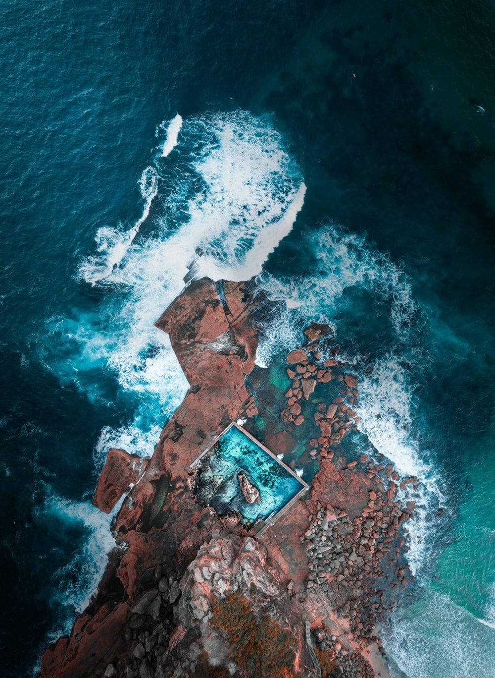 海洋 桌布 壁紙 — 從高空俯瞰，一個孤獨的人漂浮在一個雕刻在紅色岩石中的方形水池中，周圍是洶湧的綠松石色海浪。