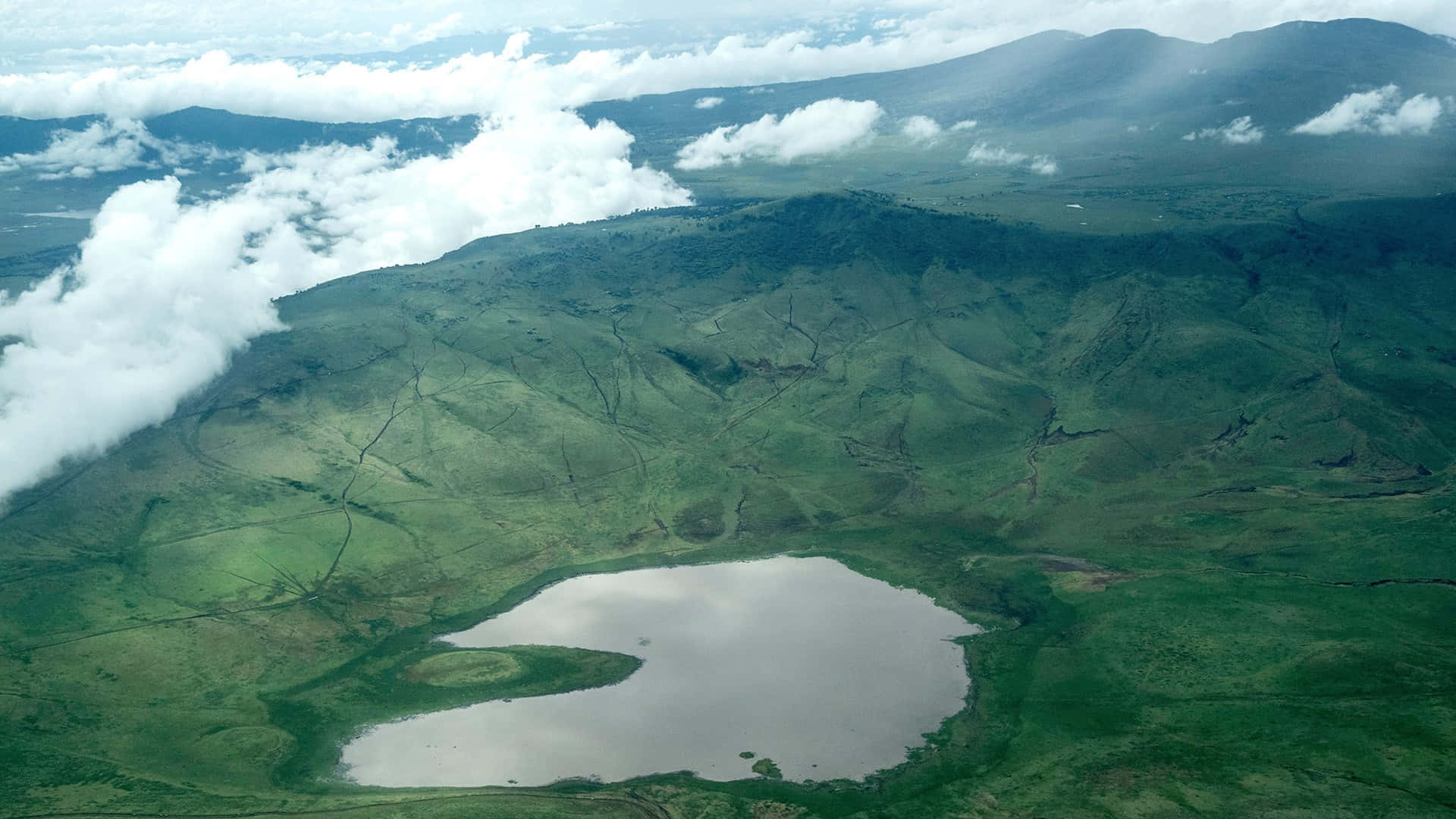 Tanzania 桌布 壁紙 — 從高空俯瞰，一個寧靜的火山口湖坐落在鬱鬱蔥蔥的青山之中，部分被飄動的白雲遮蓋。