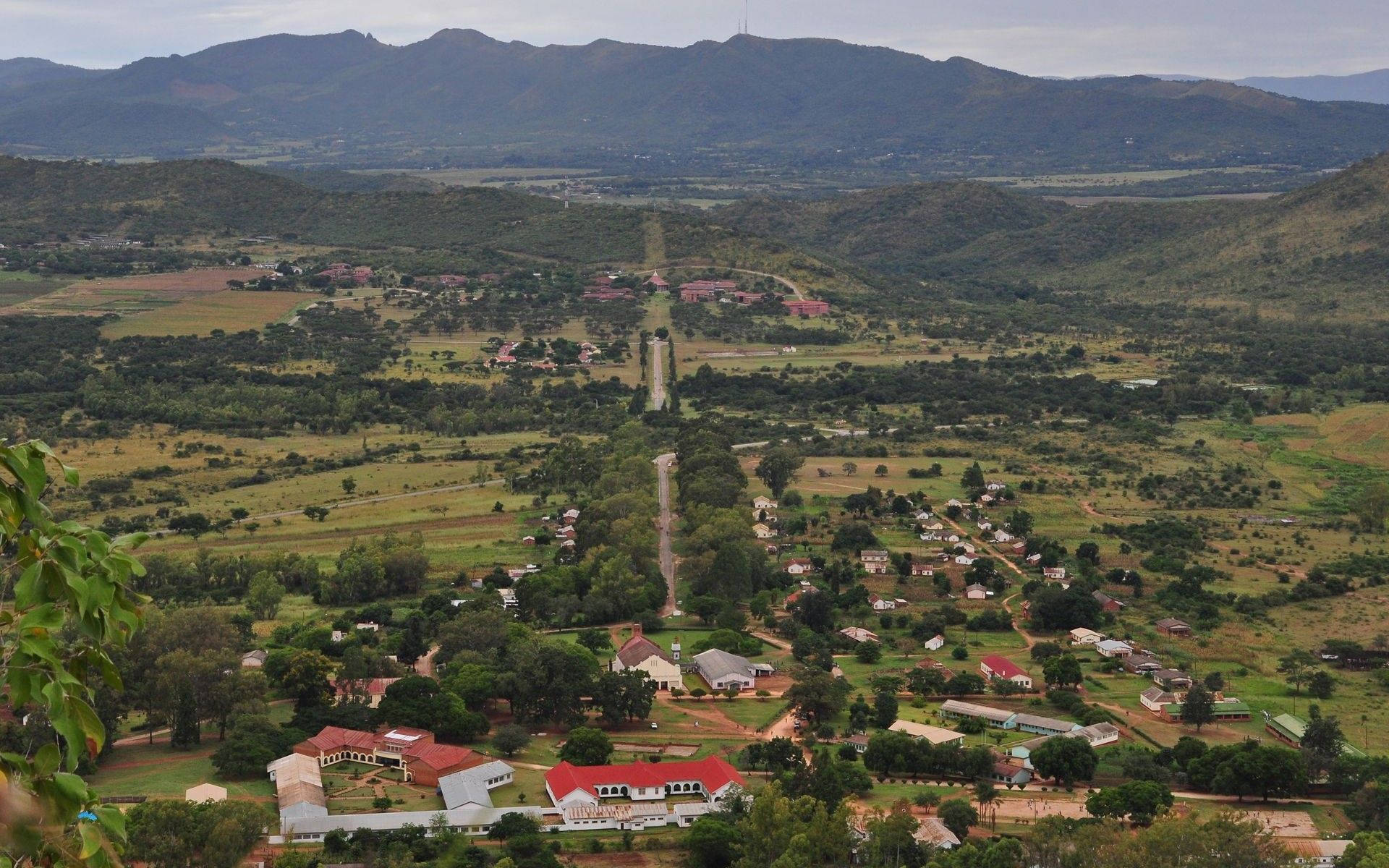 Zimbabwe 桌布 壁紙 — 從高空俯瞰，一個廣闊的山谷小鎮坐落在連綿起伏的綠色山丘之間，遠處有山脈，天空多雲。
