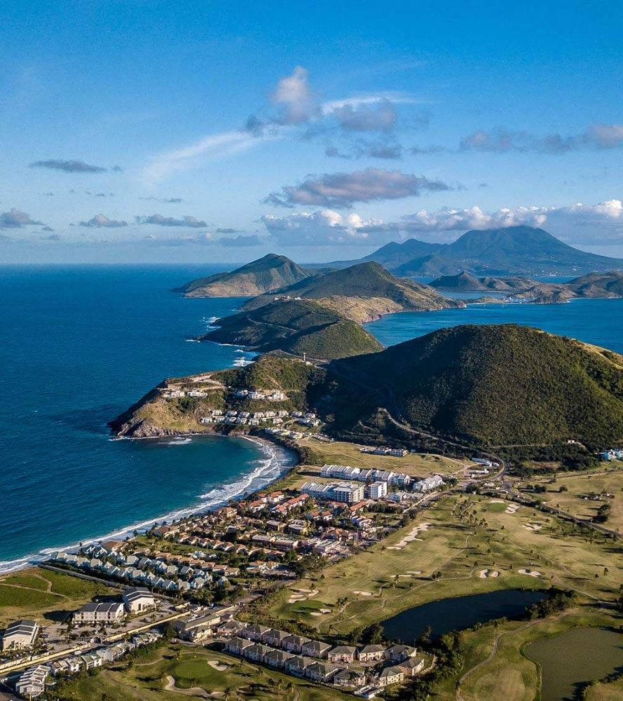 島嶼 桌布 壁紙 — 從高空俯瞰，一個沿海小鎮坐落在碧綠海水與蒼翠山巒之間，上方是明亮的藍天。