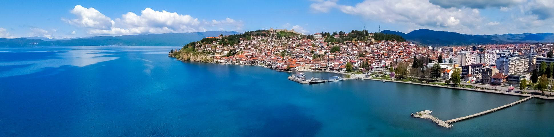 Macedonia 桌布 壁紙 — 從高空俯瞰，一個色彩繽紛的沿海小鎮坐落在山坡上，俯瞰著廣闊的深藍色湖泊，天空明亮，雲朵蓬鬆。