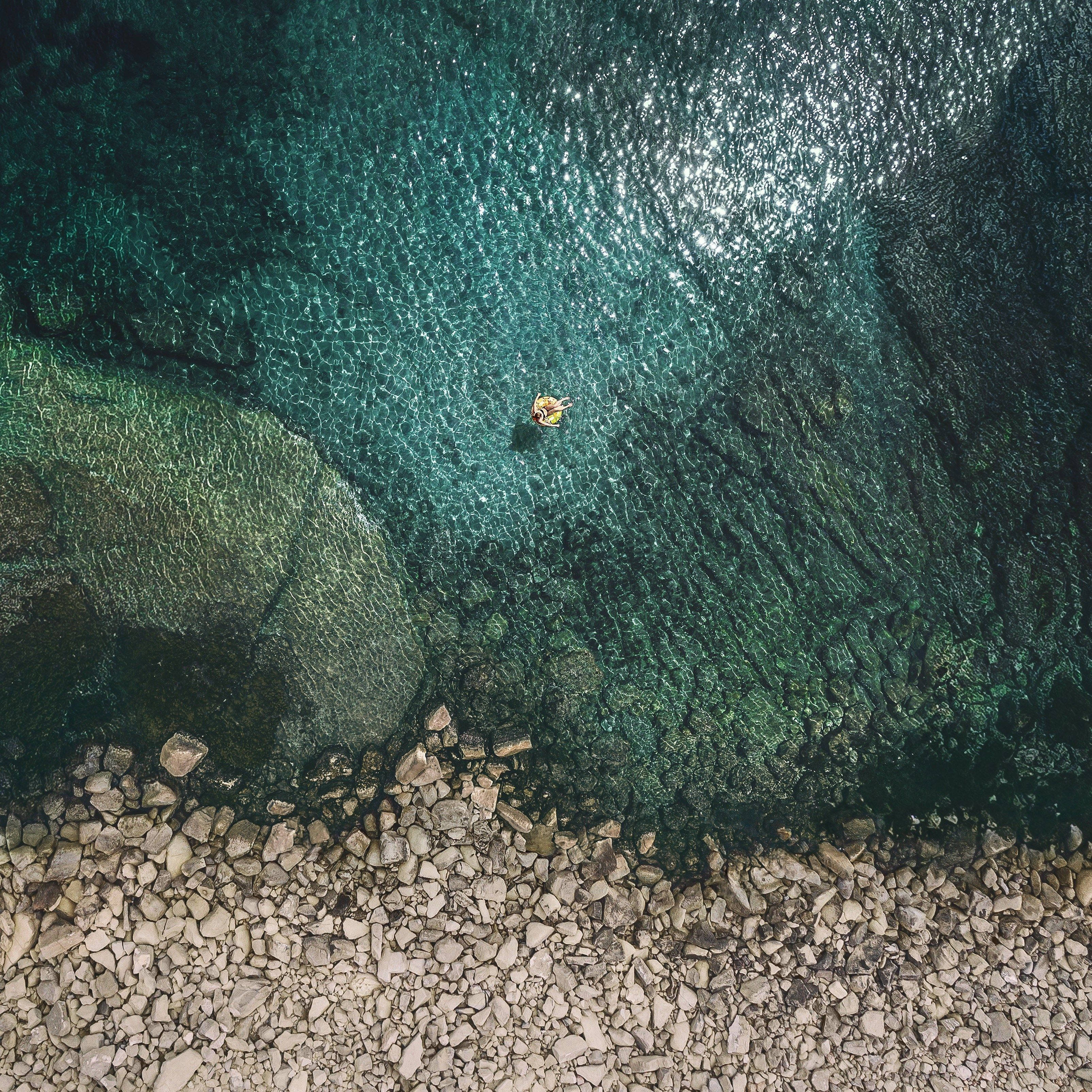 iPad 桌布 壁紙 — 從高空俯瞰，一名孤獨的泳者漂浮在清澈碧綠的海水中，旁邊是岩石海岸線。
