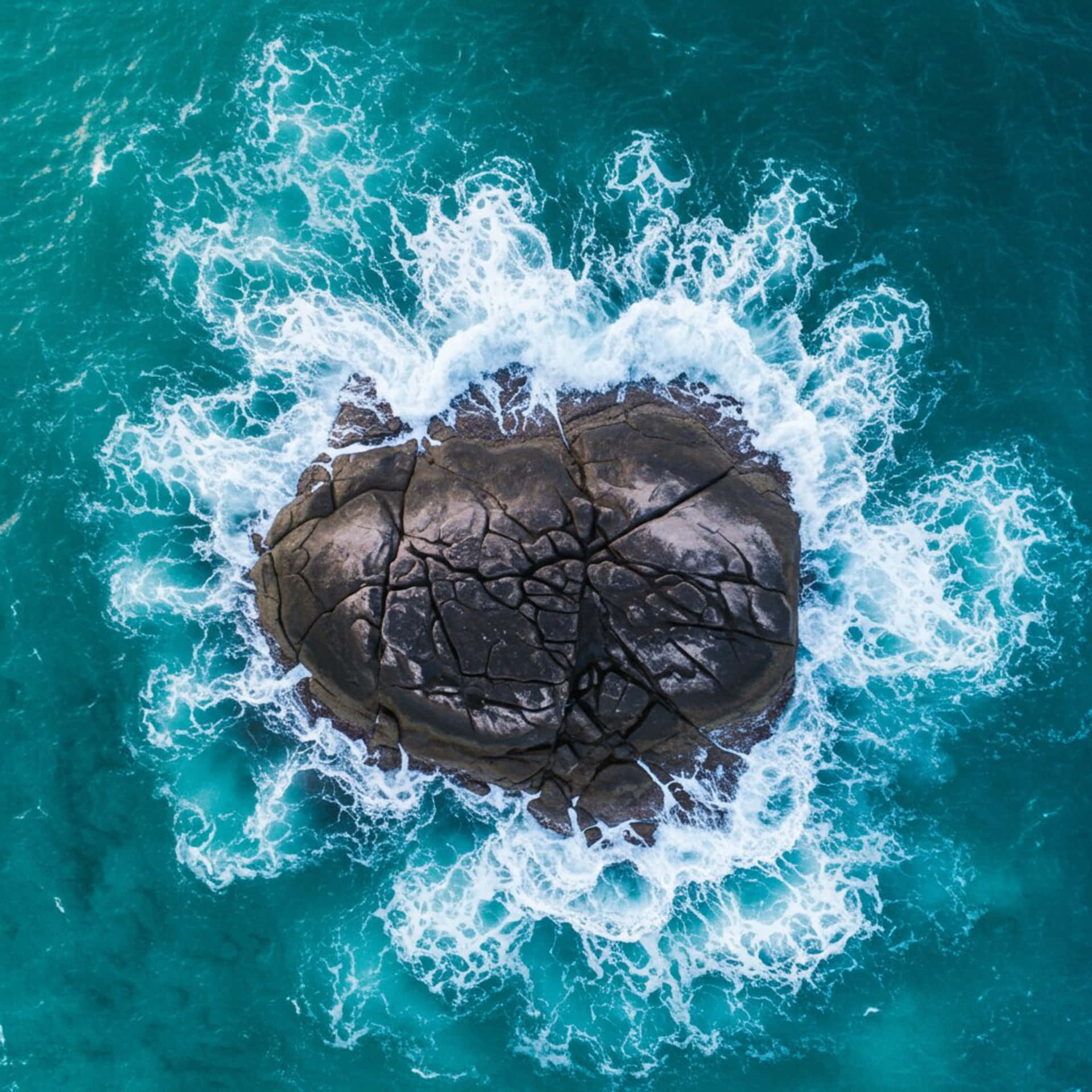 Island on the sea 壁紙 — 從高空俯瞰，一塊深色裂開的岩石被碧綠色的海浪拍打環繞。