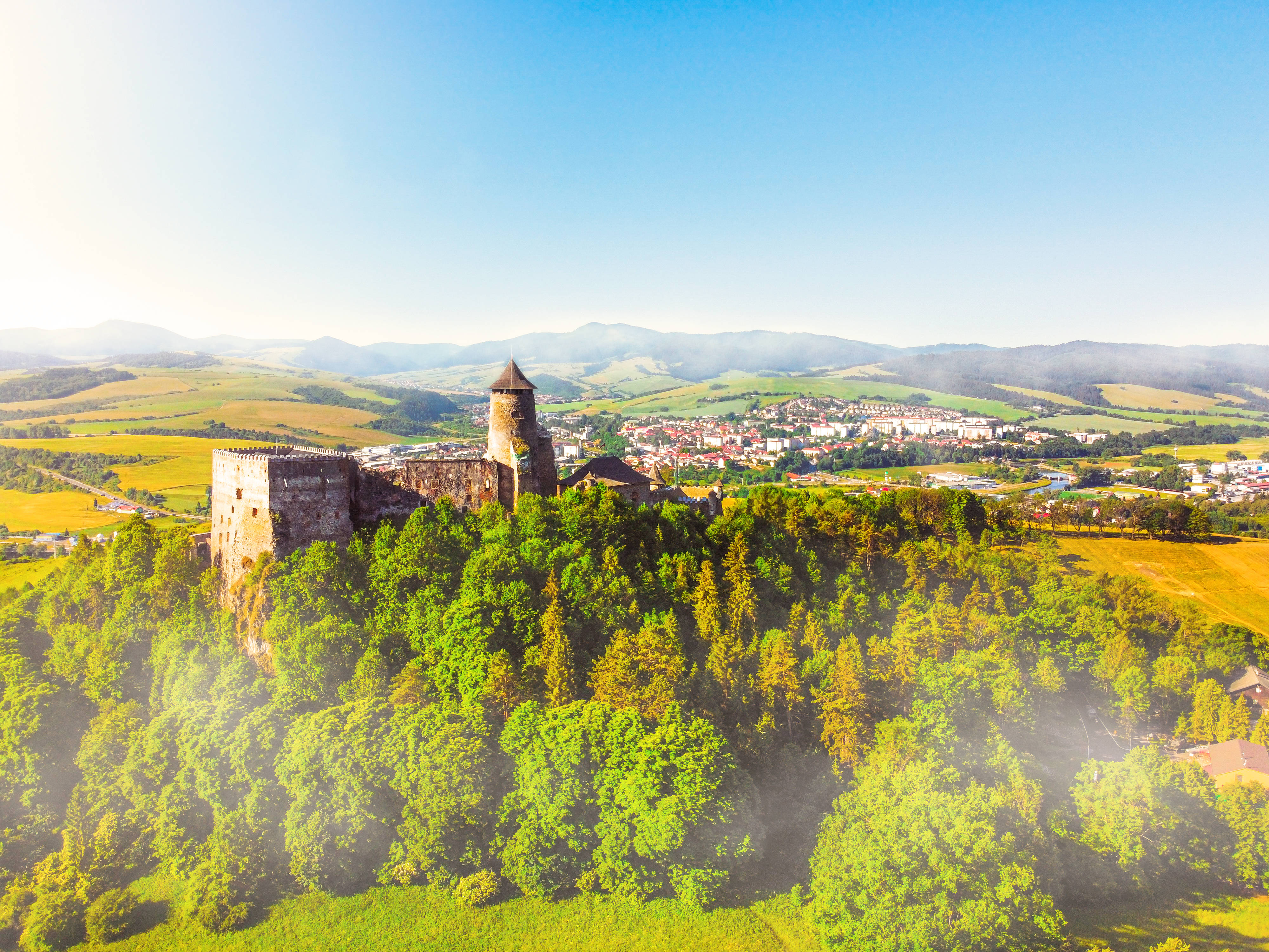 Slovakia 桌布 壁紙 — 從高空俯瞰，一座中世紀城堡坐落在鬱鬱蔥蔥的綠色山丘上，俯瞰著山谷小鎮，天空湛藍清澈。