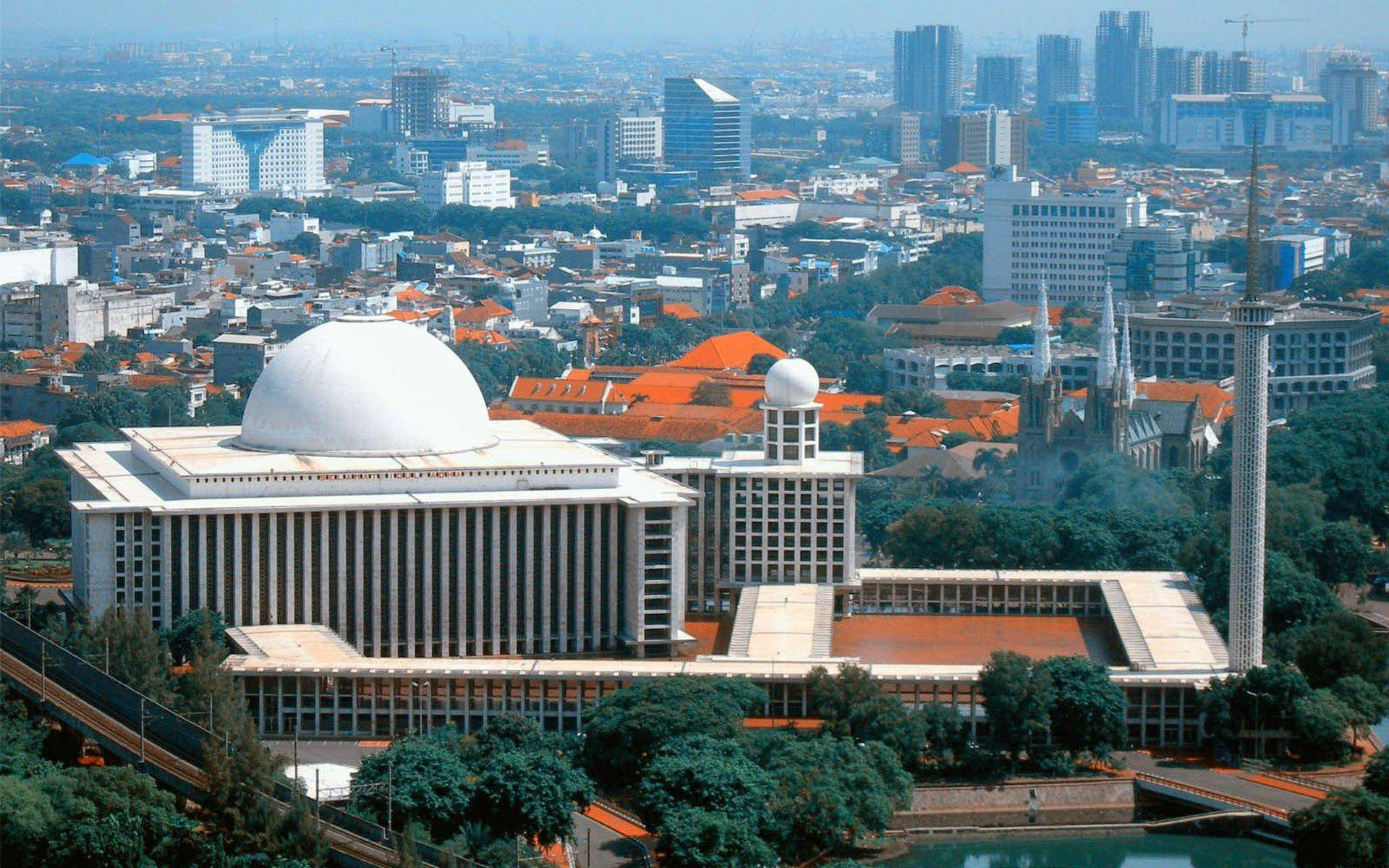 Jakarta 桌布 壁紙 — 從高空俯瞰，一座宏偉的白色圓頂清真寺，帶有宣禮塔，矗立在密集的現代摩天大樓和紅頂建築組成的城市叢林中，籠罩在朦朧的日光下。