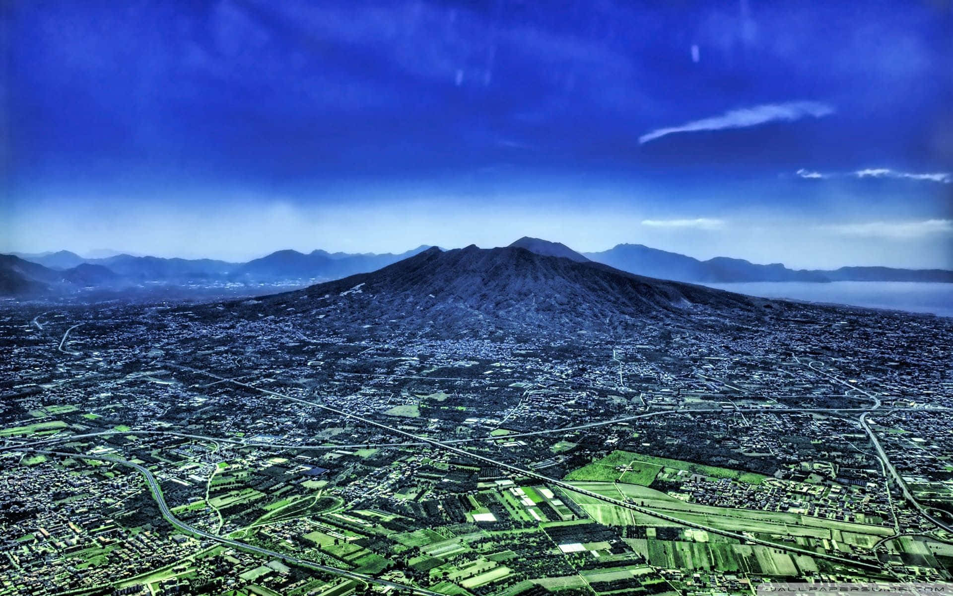 Volcano 桌布 壁紙 — 從高空俯瞰，一座廣闊的城市坐落於巨大火山腳下，天空蔚藍壯麗。