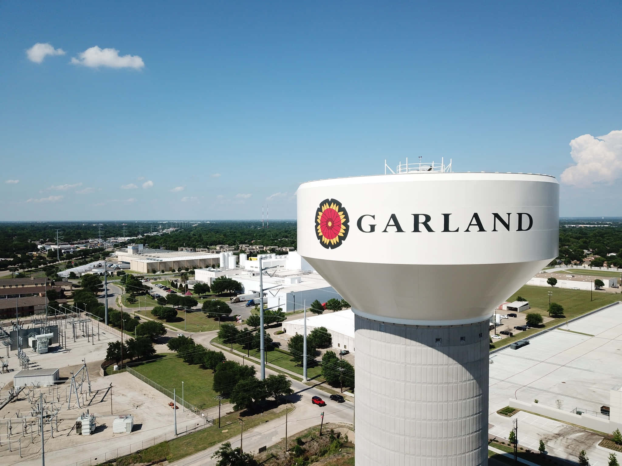 德州 桌布 壁紙 — 從高空俯瞰，一座白色水塔上印有「GARLAND」和向日葵標誌，在晴朗藍天下主導著郊區景觀。