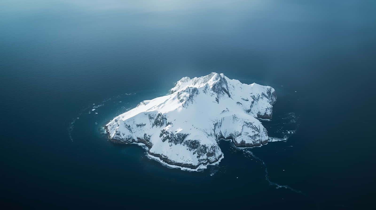 Majestic Sharp Snowy Mountain Peak Island Wallpaper 壁紙 — 從高空俯瞰，一座白雪覆蓋的山島從深藍色海洋中陡峭升起。