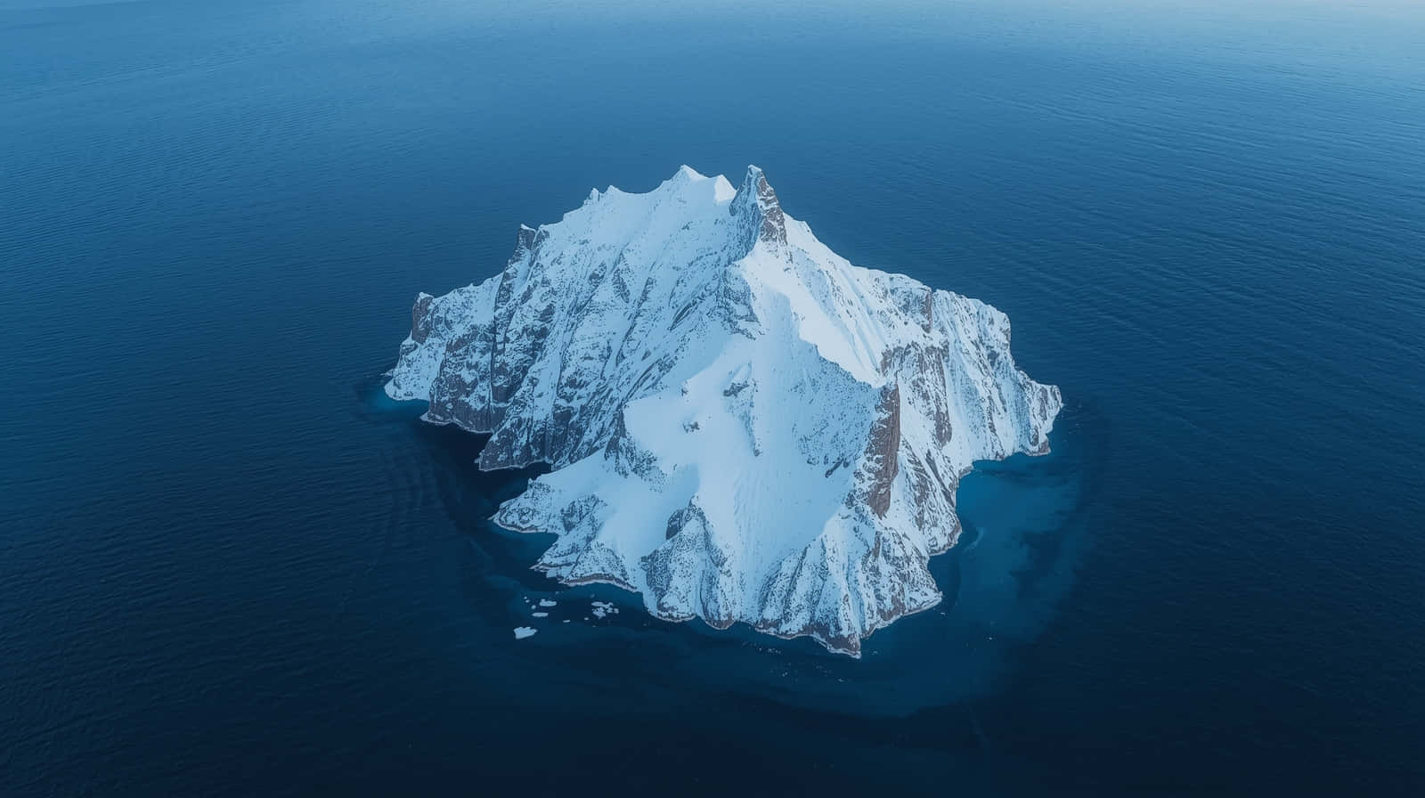 Aerial View of Snow Covered Arctic Island Wallpaper 壁紙 — 從高空俯瞰，一座白雪覆蓋的岩石島嶼從深藍色海洋中陡峭升起。