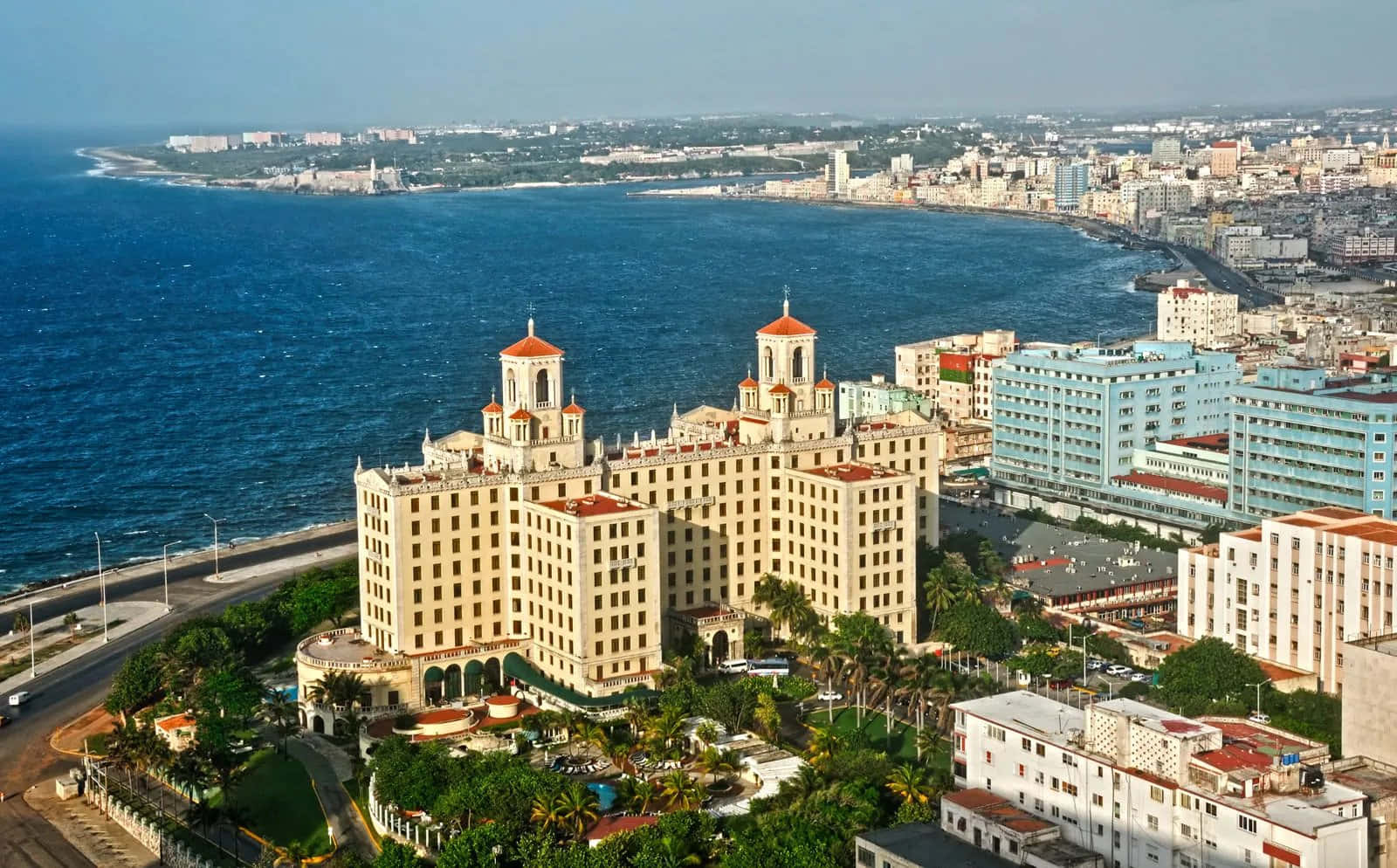 Cuba 桌布 壁紙 — 從高空俯瞰，一座米色大酒店，紅色屋頂，隱藏在棕櫚樹之間，旁邊是湛藍的海洋和廣闊的海濱城市。