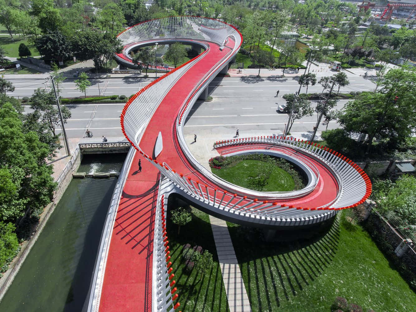 bridge 桌布 壁紙 — 從高空俯瞰，一條引人注目的紅白相間蛇形行人天橋蜿蜒穿過翠綠城市公園中的運河和道路。