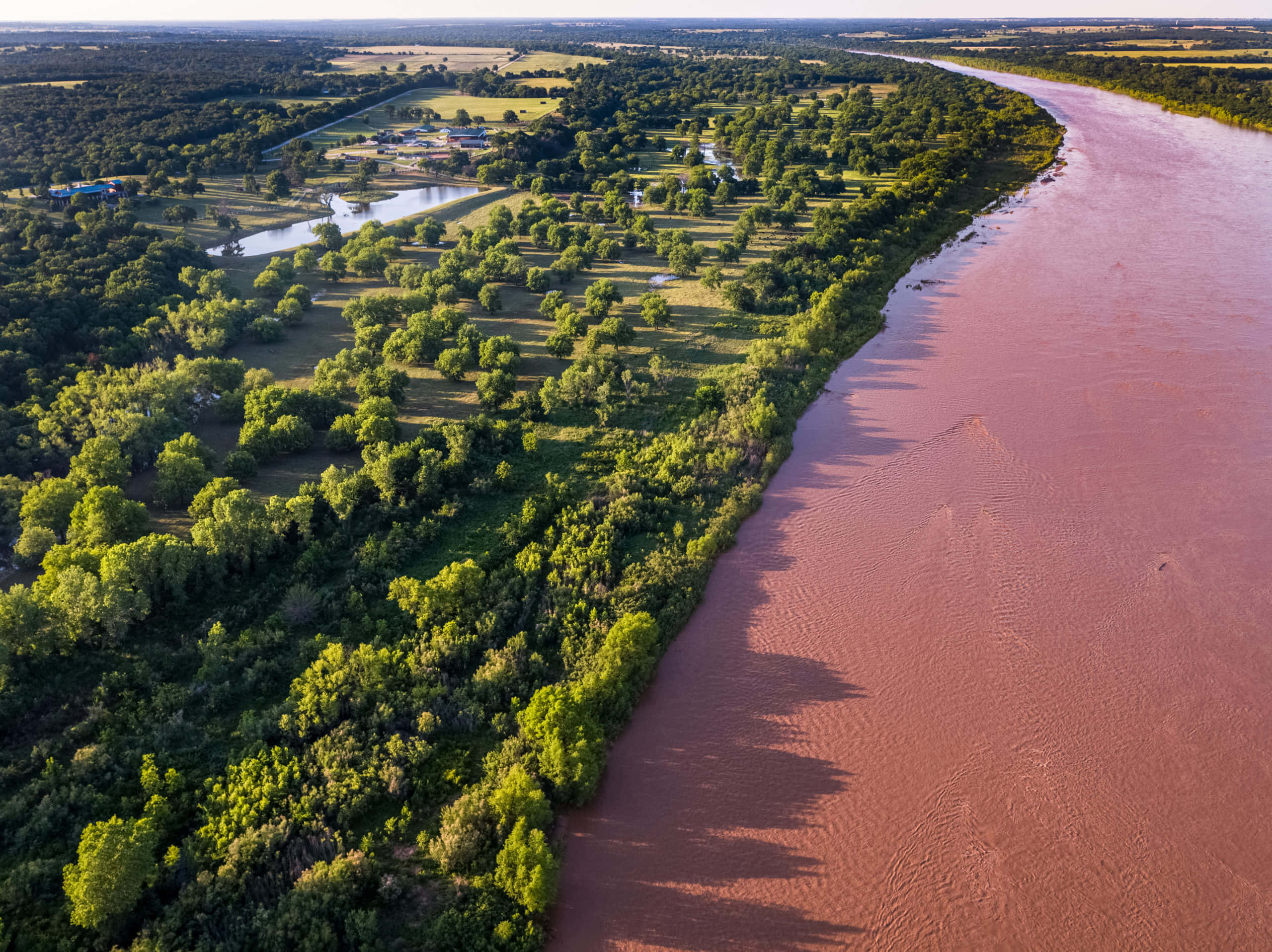 river 桌布 壁紙 — 從高空俯瞰，一條蜿蜒的泥濘河流在明媚的日光下穿過茂密的綠色森林和農田。