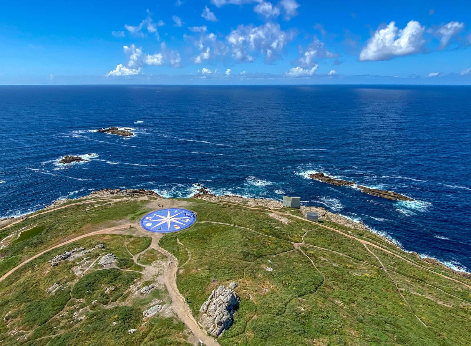 島嶼 桌布 壁紙 — 從高空俯瞰，一片草叢生的海岸懸崖上，有一個大型藍色指南針玫瑰藝術裝置，俯瞰著深藍色的海洋和散落的岩石，天空明亮。