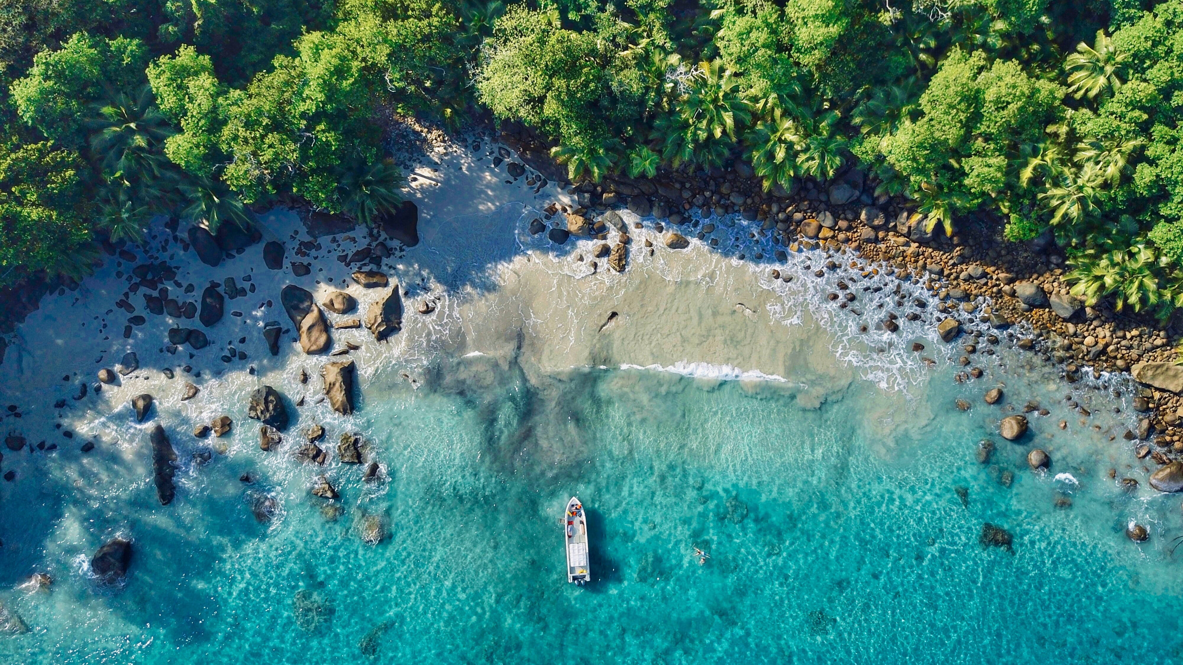 島嶼 桌布 壁紙 — 從高空俯瞰，一艘孤獨的小船漂浮在清澈碧綠的海水中，旁邊是岩石密布的熱帶海灘，長滿了茂密的綠色棕櫚樹。