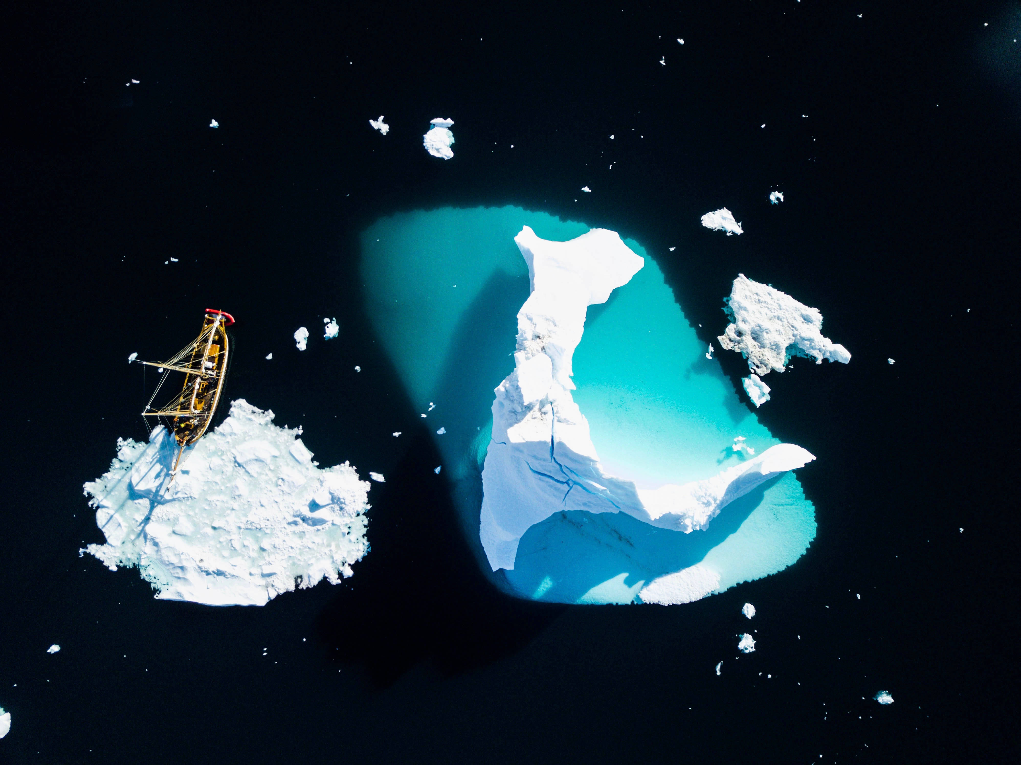 Greenland 桌布 壁紙 — 從高空俯瞰，一艘小船停泊在白雪覆蓋的冰山上，旁邊是巨大的藍綠色浮冰，漂浮在黑暗的海水中。
