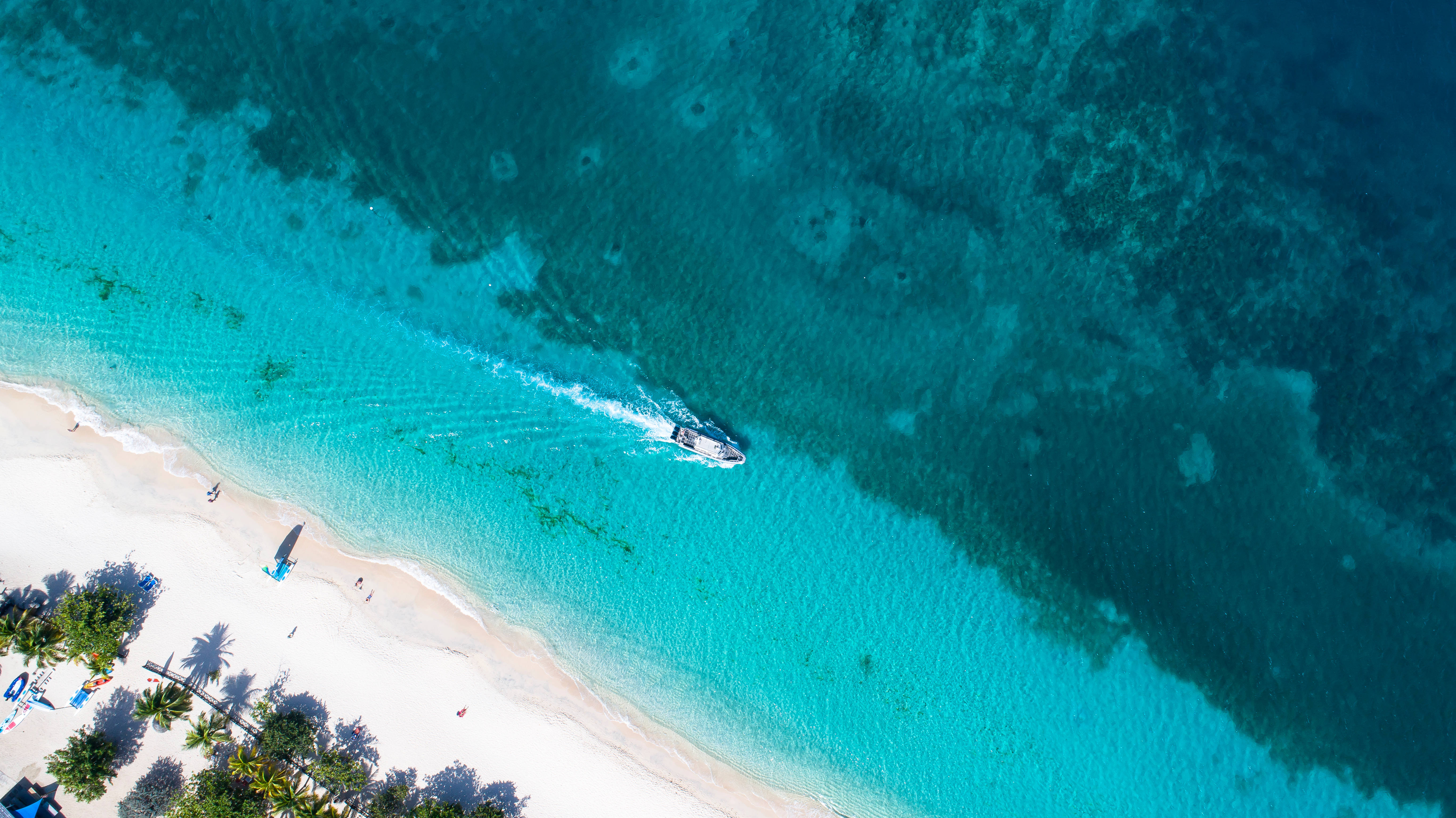 Grenada 桌布 壁紙 — 從高空俯瞰，一艘快艇在碧綠色的海面上飛馳，旁邊是種滿棕櫚樹的白色沙灘。