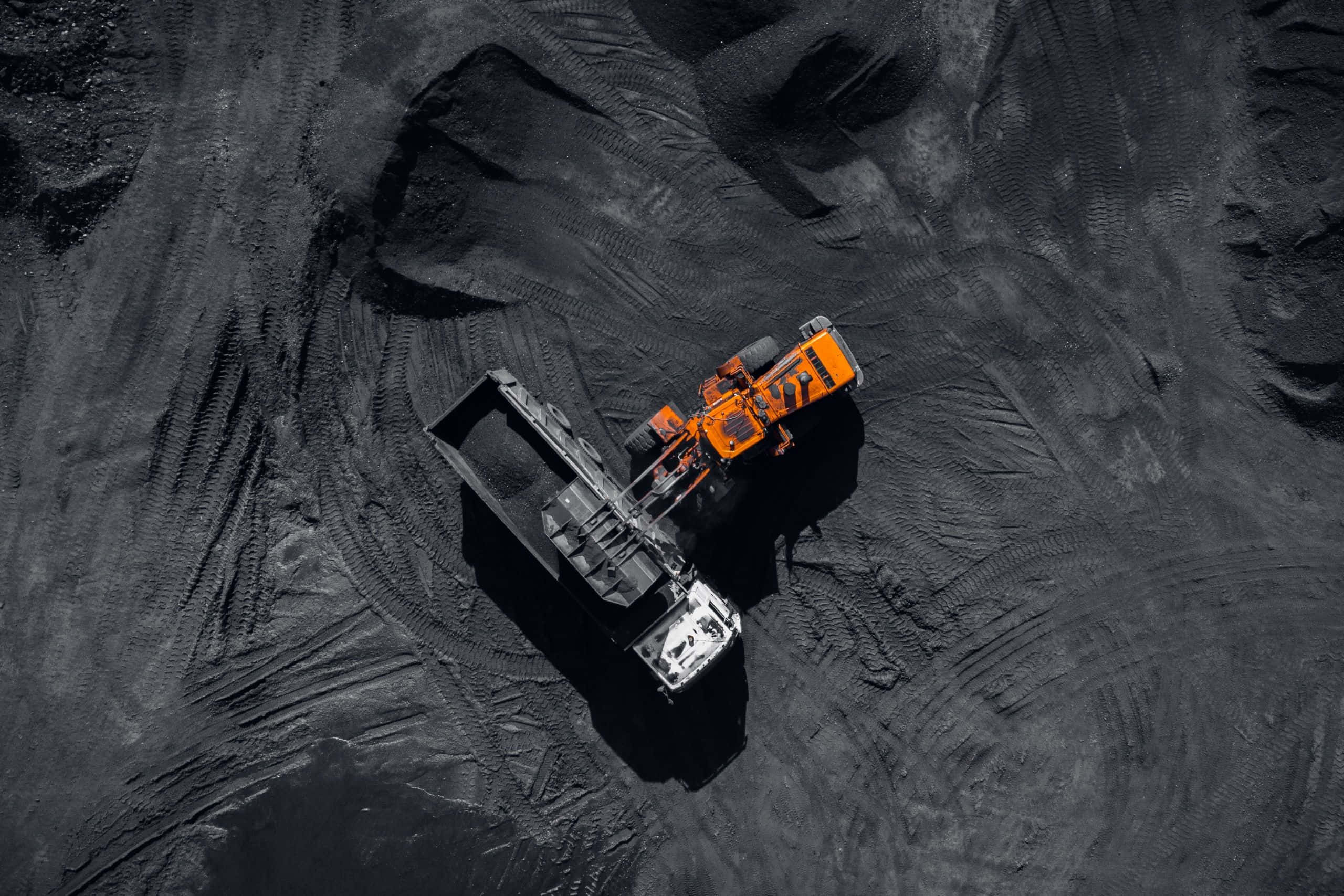 Space Mining 桌布 壁紙 — 從高空俯瞰，一輛橙色裝載機正在廣闊的煤堆上將煤炭卸入黑色自卸卡車。
