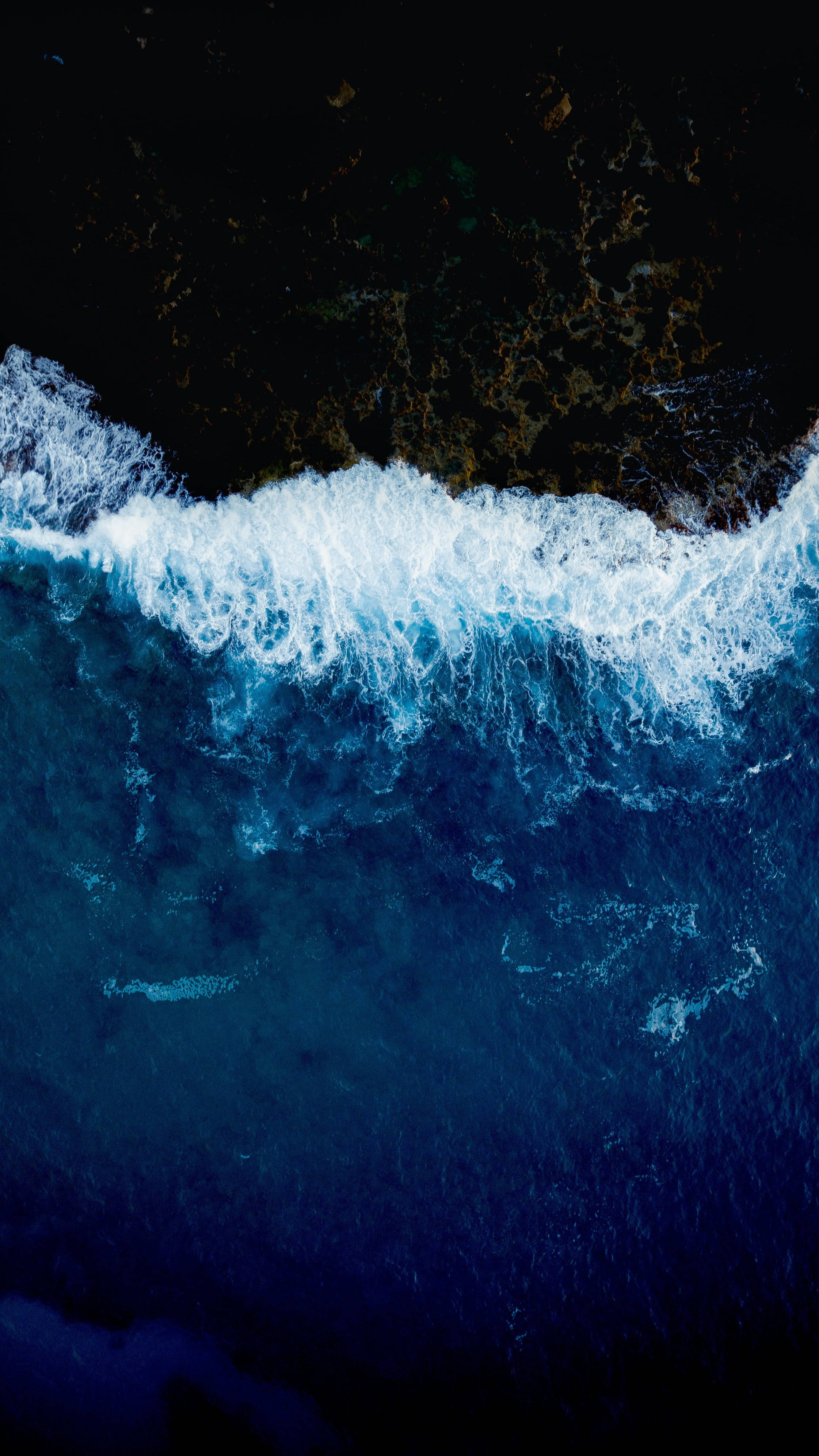 Wave 桌布 壁紙 — 從高空俯瞰，一道強勁的海浪拍打著黑暗的岩石海岸，激起鮮豔的藍色和白色泡沫。