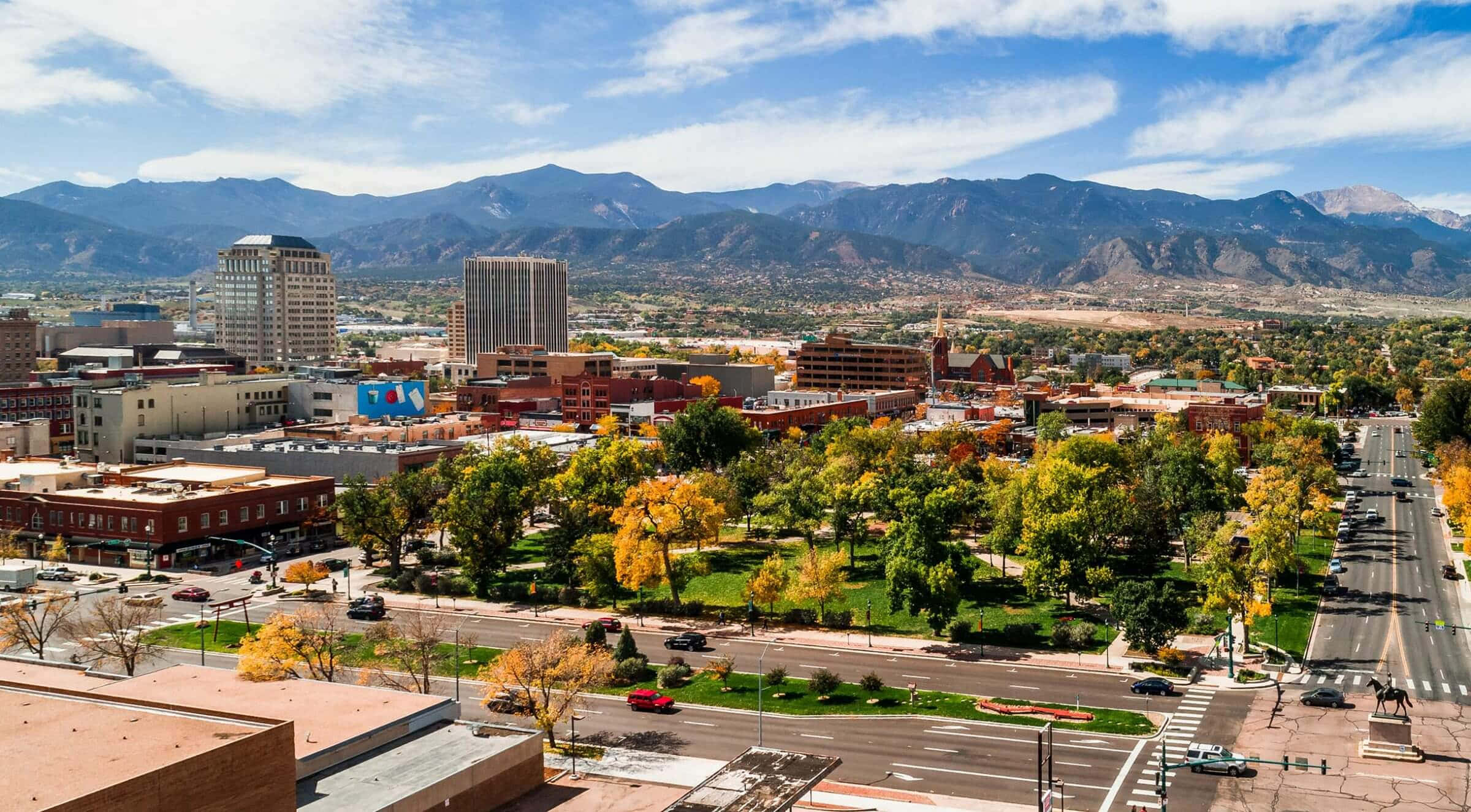 Colorado 桌布 壁紙 — 從高空俯瞰，充滿活力的城市景觀坐落在城市建築和秋色樹木之間，背景是雄偉的山脈，天空湛藍明亮。