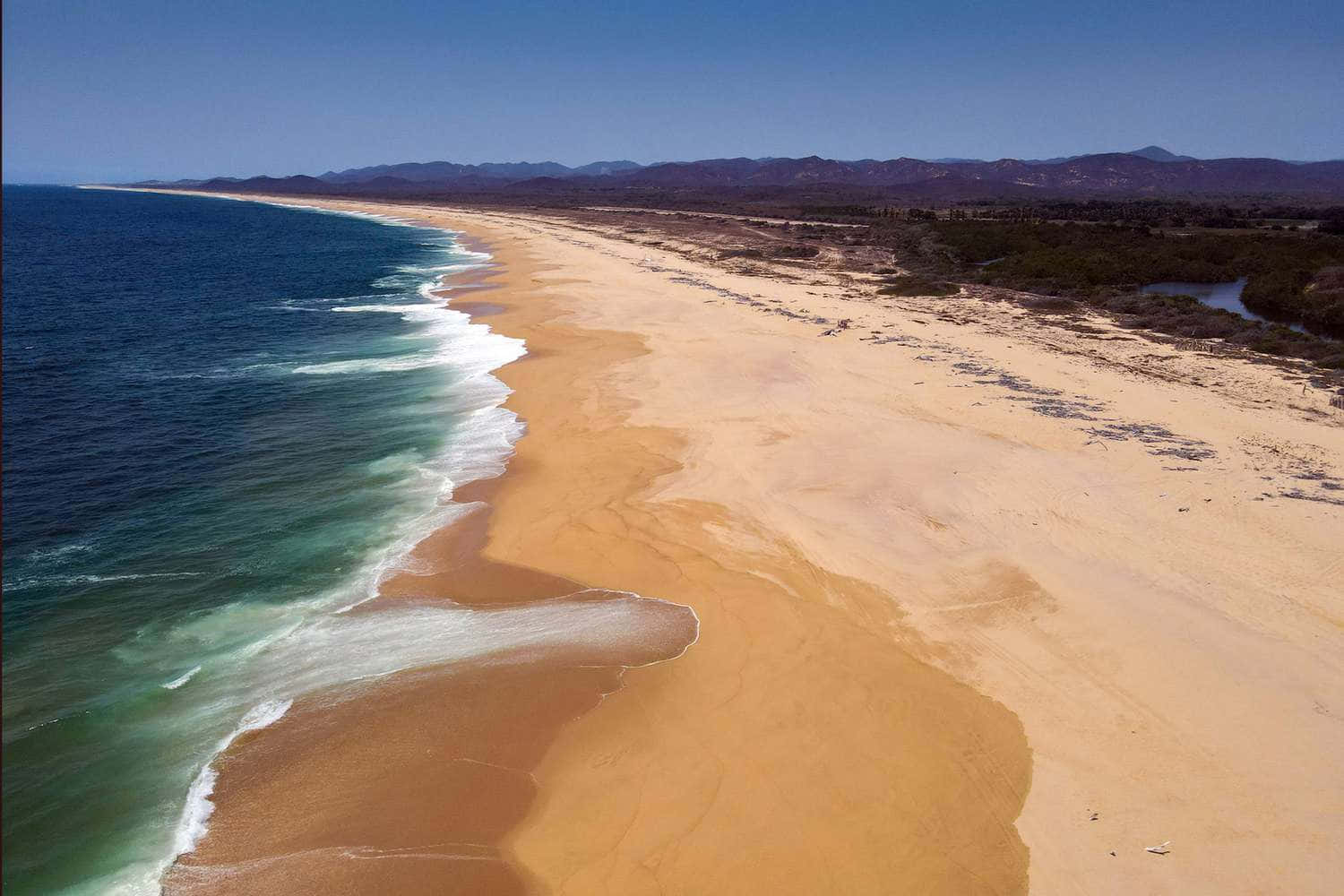 Mexican Beach 桌布 壁紙 — 從高空俯瞰，在清澈藍天下，一條長長的金色沙灘沿著碧綠的海浪蜿蜒伸展。