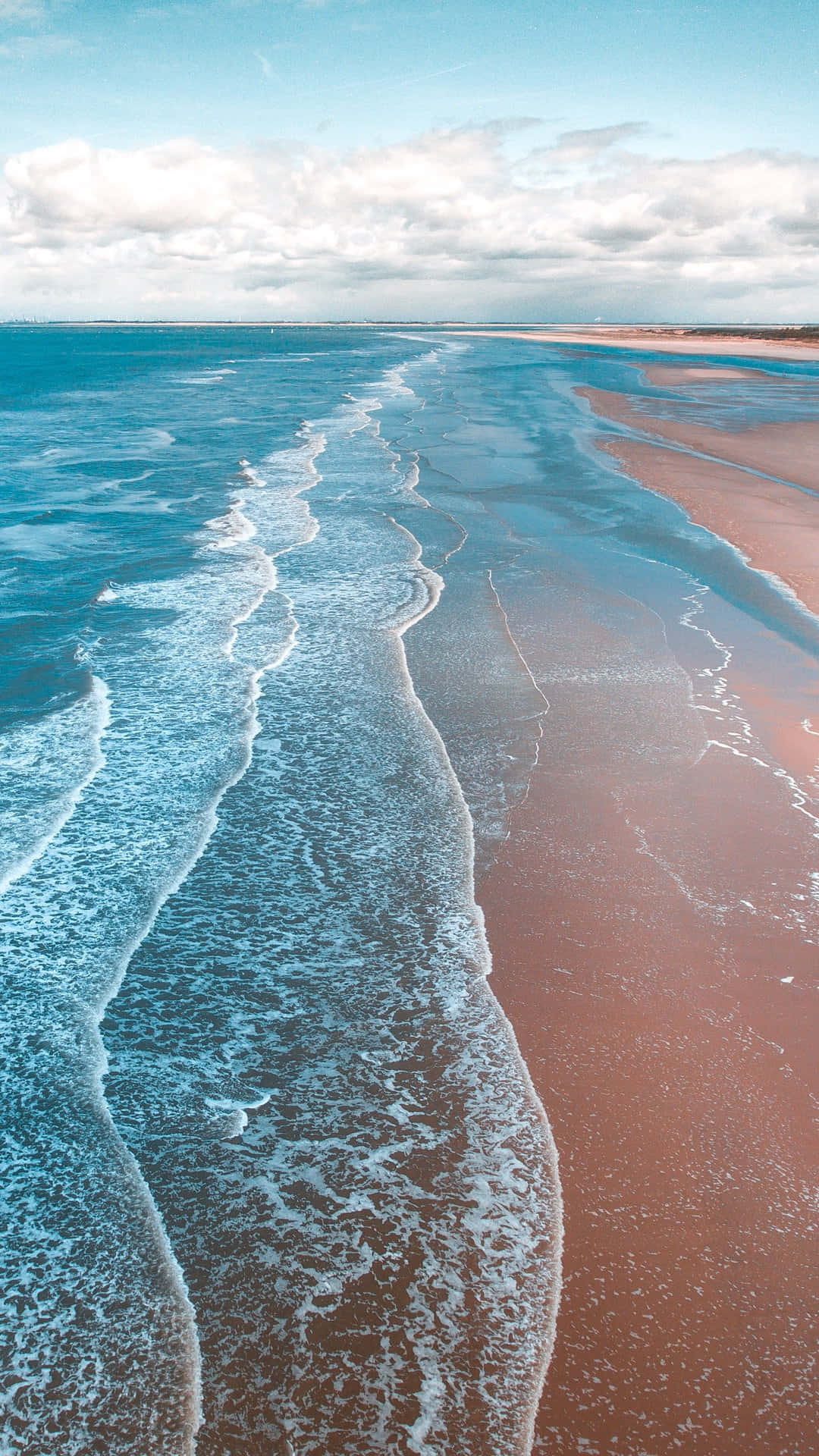 Surfing iPhone 桌布 壁紙 — 從高空俯瞰，在部分多雲的天空下，碧綠色的海浪拍打著粉紅色的沙灘。