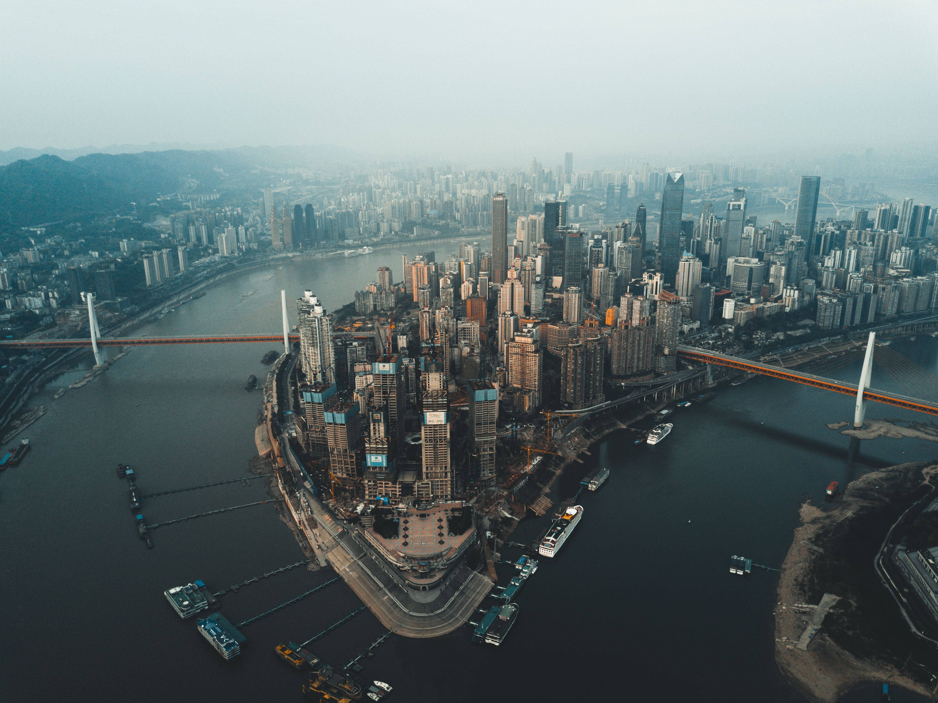 Chongqing 桌布 壁紙 — 從高空俯瞰，密集的現代城市景觀沿著蜿蜒的河流分佈，橋樑和船隻在朦朧的天空下。