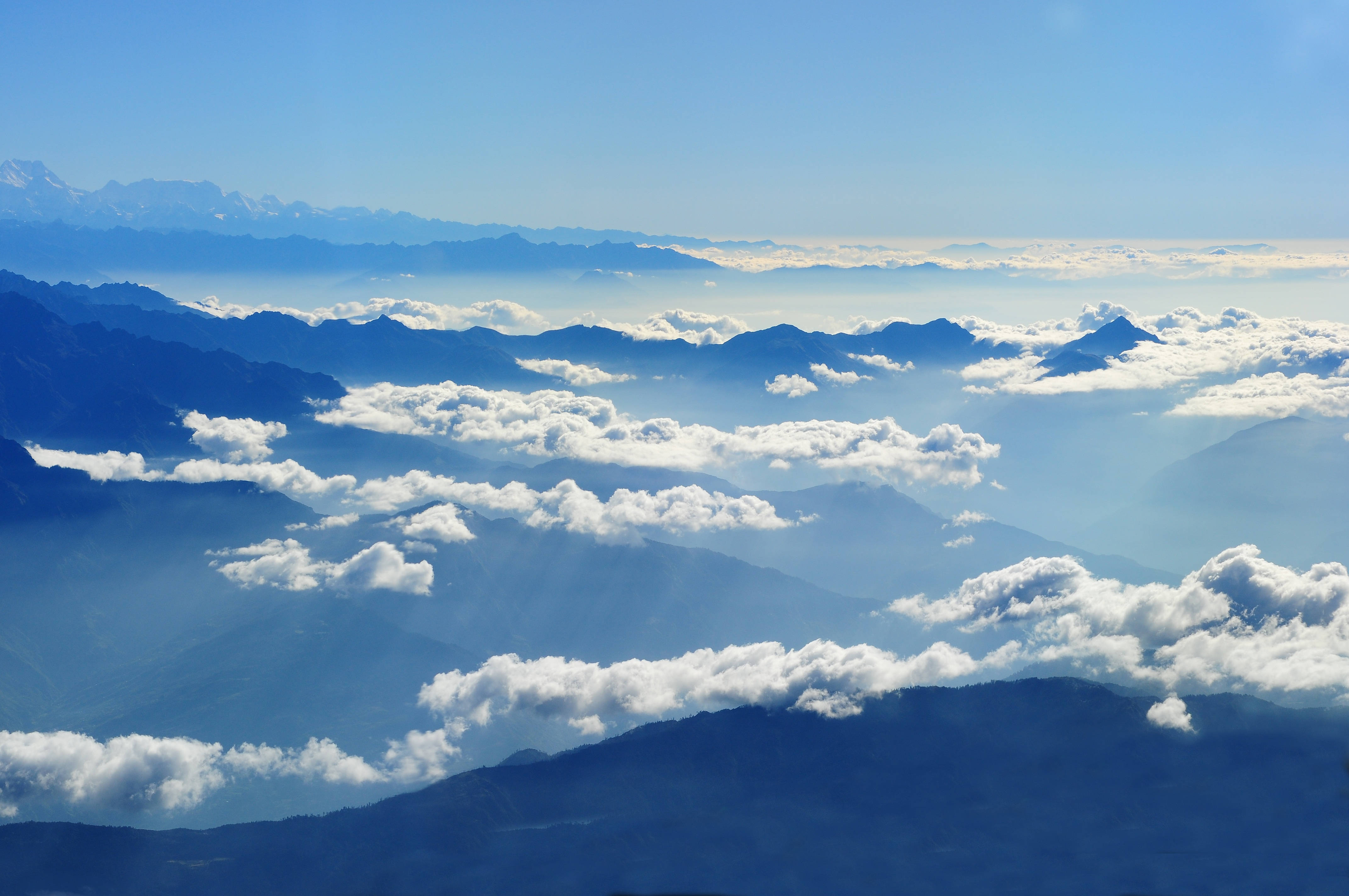 天空 桌布 壁紙 — 從高空俯瞰，層層疊疊的山脈穿透蓬鬆的白雲，在晴朗的藍天下，陽光穿透雲層。