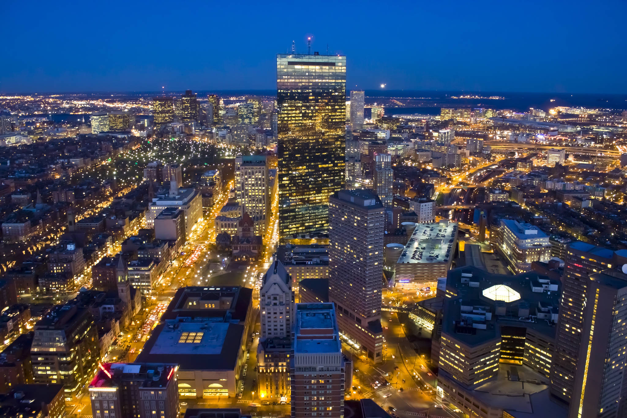 Boston 桌布 壁紙 — 從高空俯瞰，暮色中廣闊的城市景觀，摩天大樓燈火通明，街道閃爍，襯托著深藍色的天空。