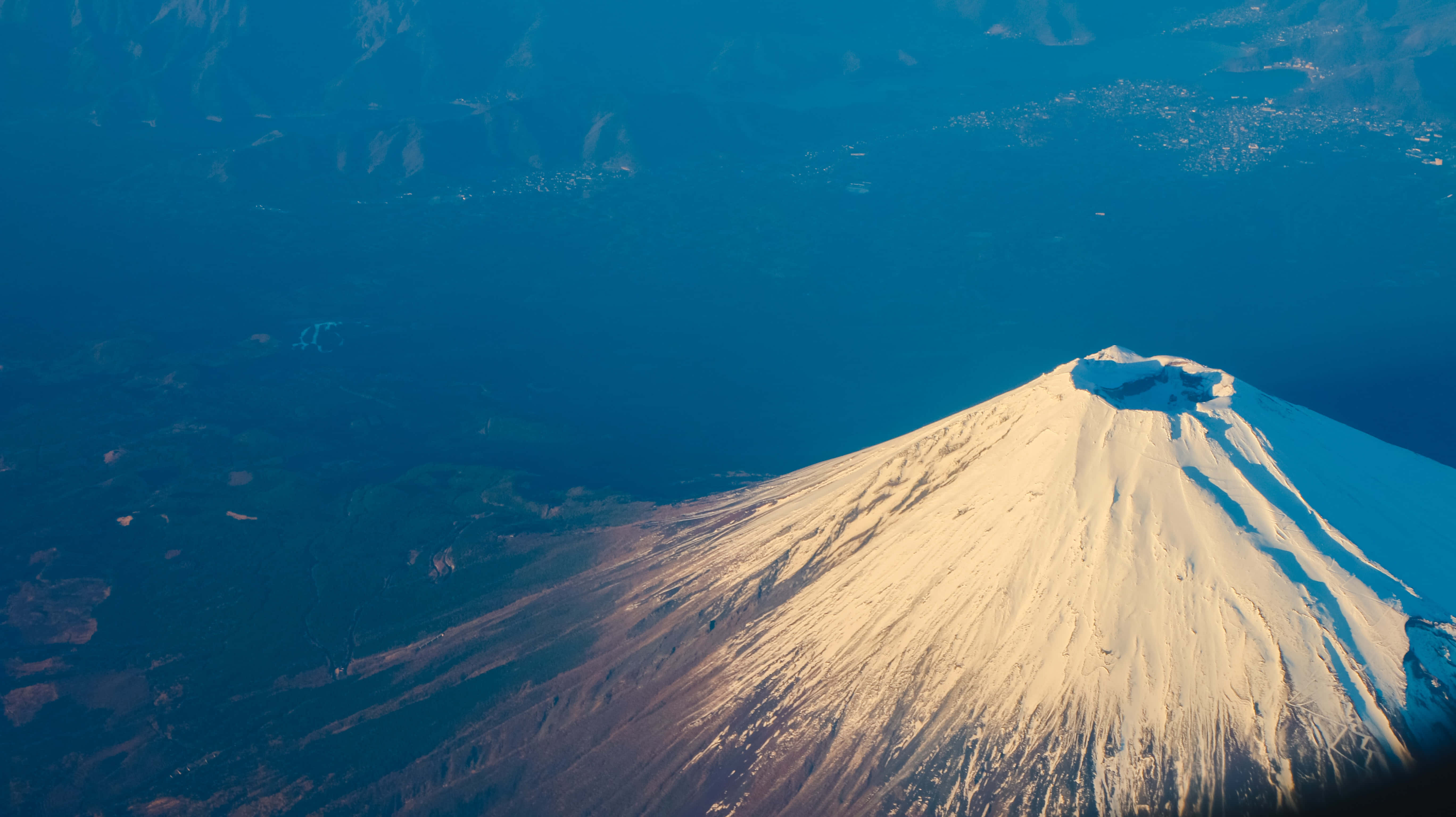 日本 桌布 壁紙 — 從高空俯瞰，白雪覆蓋的富士山沐浴在金色陽光下，背景是深藍色的海洋和森林覆蓋的山坡。