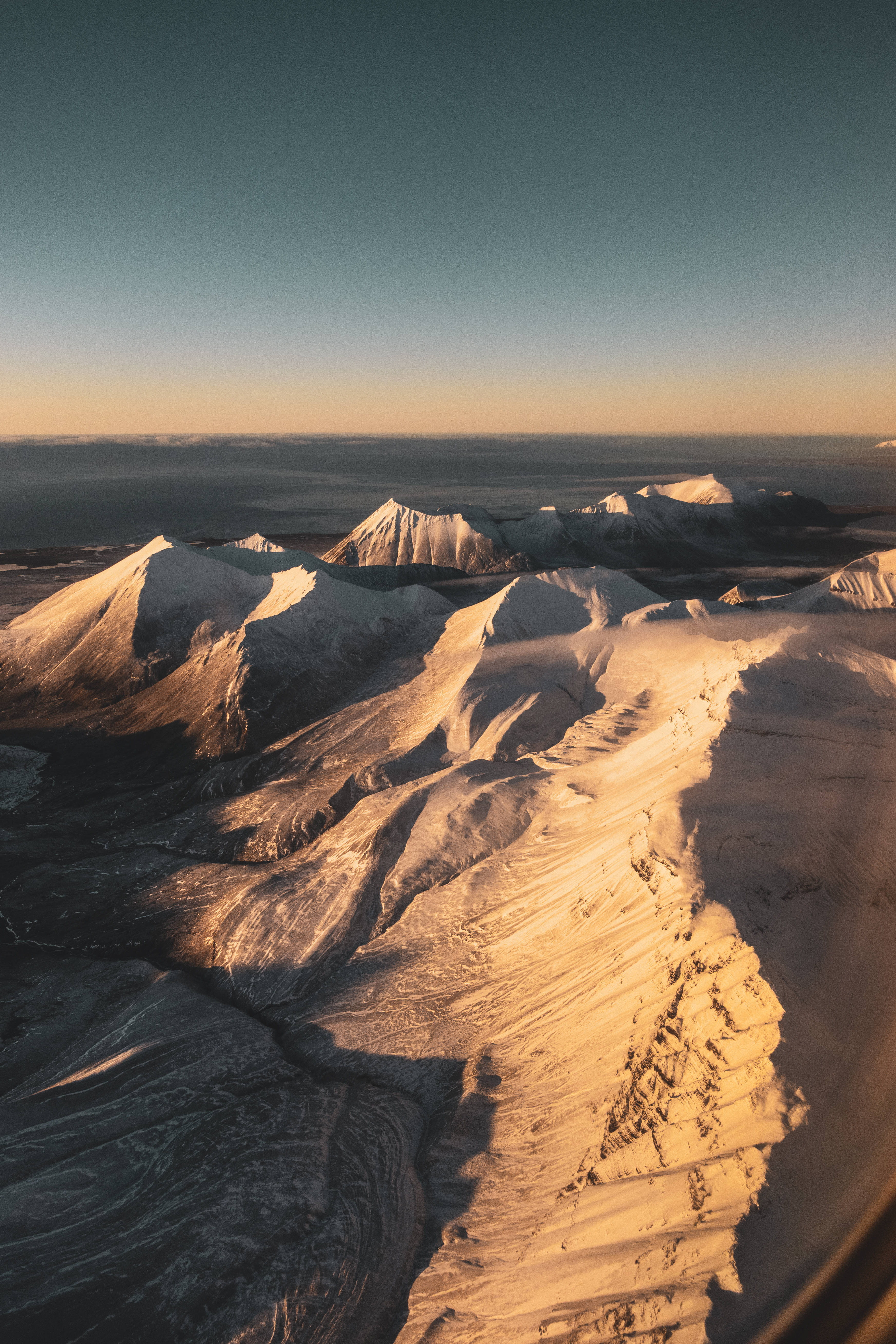Svalbard 桌布 壁紙 — 從高空俯瞰，白雪覆蓋的山峰沐浴在金色日出光芒中，襯托著深藍色的天空。